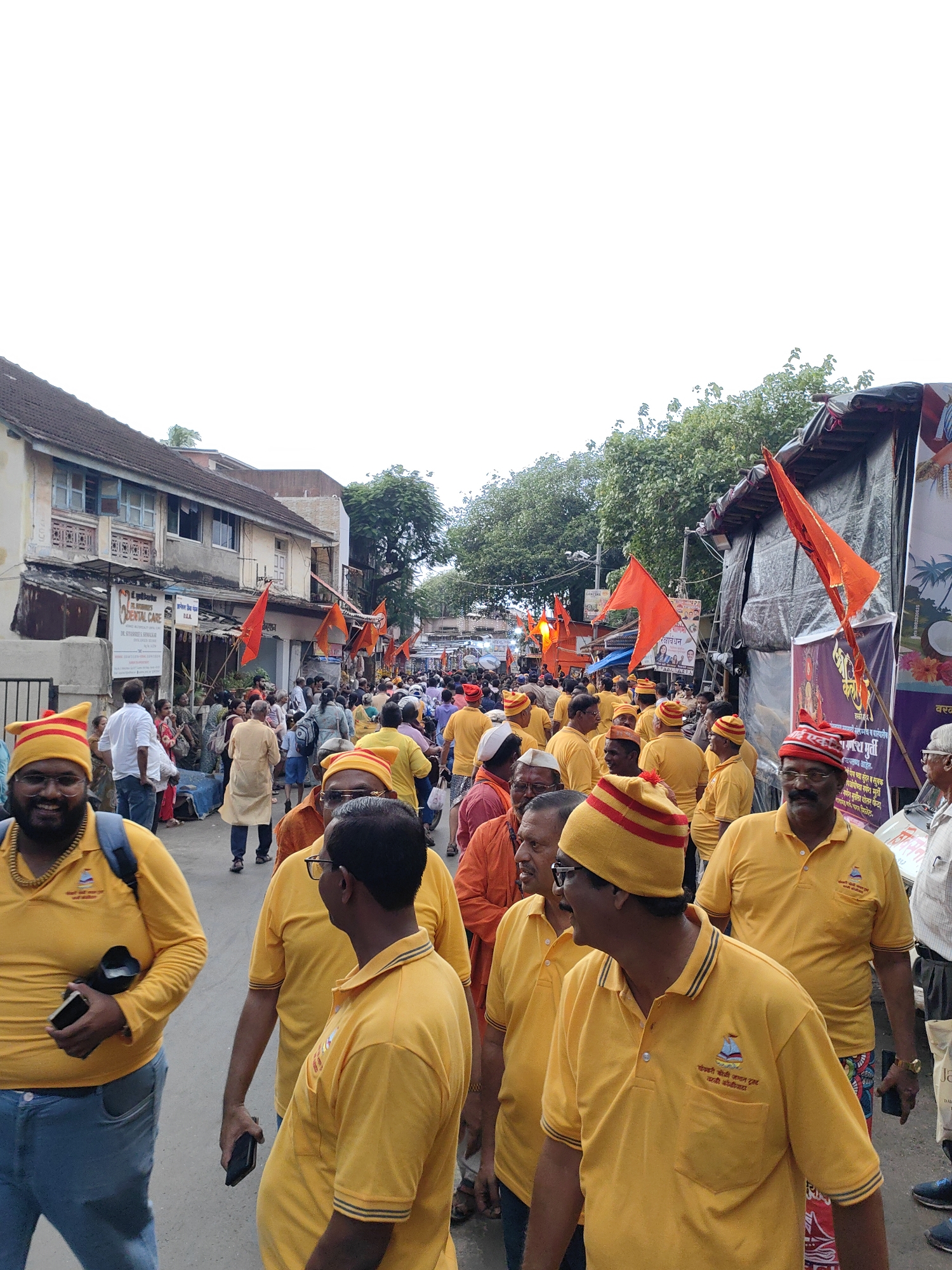 A crowd of people are walking down a street lined with buildings and trees. The people are mostly wearing yellow shirts and many are wearing yellow and red striped hats. Some people are holding orange flags. The buildings appear to be in a commercial area, with some shops visible. The overall scene suggests a religious procession or gathering. The presence of a dental care sign in English suggests an area with a mix of local and potentially international influence. The architecture and vegetation are consistent with a city in India, possibly Mumbai, given the common use of yellow in religious processions and the style of the buildings. The sky is overcast.