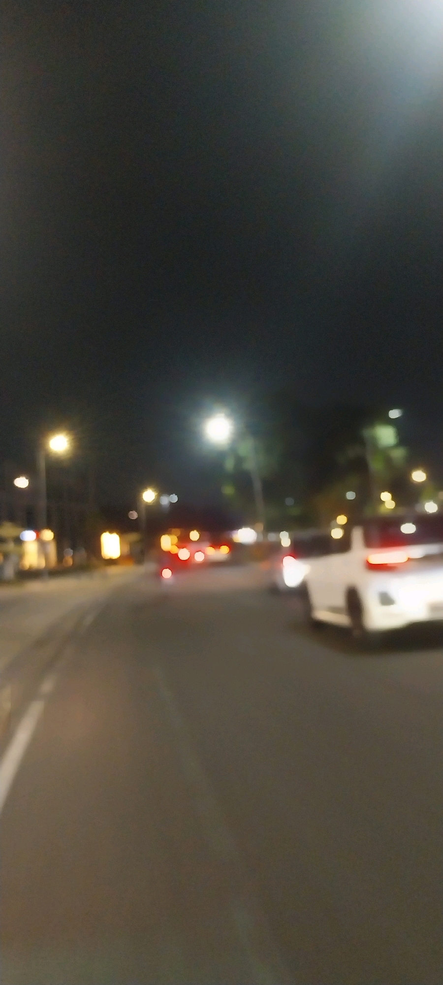The image depicts a blurry nighttime street scene in the city of Medan, Indonesia. The scene shows a road with streetlights and a few vehicles. On the right side of the image, a white car with its taillights illuminated is visible. Further down the road, the red taillights of other vehicles can be seen. Streetlights line the road, casting a glow on the dark surroundings. The overall impression is one of a typical urban environment at night, with traffic and illumination from artificial sources.