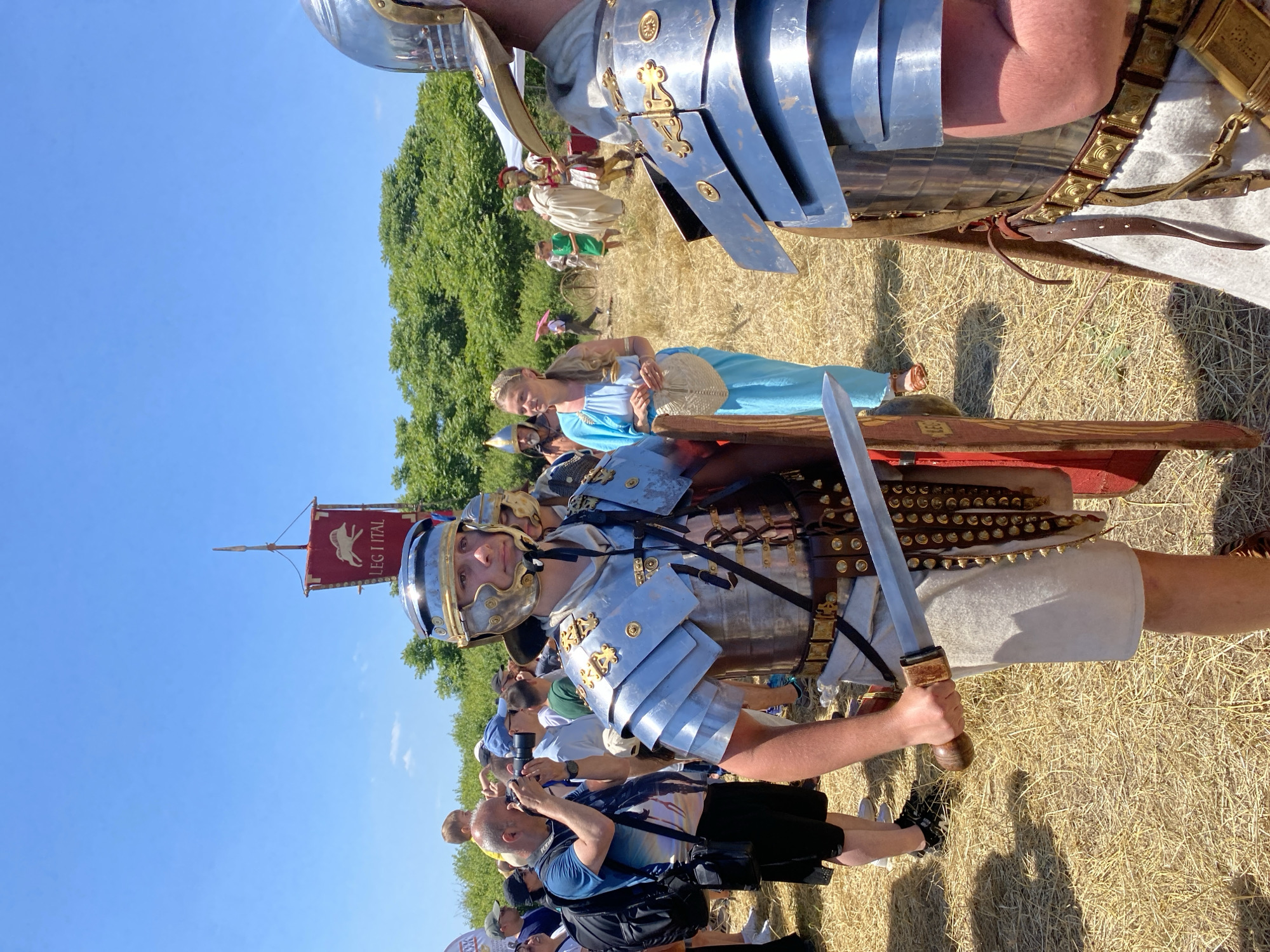 The image depicts an outdoor historical reenactment event in Gigen, Bulgaria, under a clear blue sky. The ground is covered in dry, golden-brown hay or stubble.
In the foreground, two individuals dressed as Roman legionaries are visible. The central figure, a young man, wears polished silver-colored lorica segmentata armor with gold accents, a matching Galea helmet, a white tunic, and a brown leather cingulum belt. He holds a Gladius (short sword) with a wooden hilt in his right hand and a red Scutum (shield) partially visible in his left. His feet are clad in brown caligae. To his immediate right, another Roman soldier's torso, right arm, and helmet are partially visible, showing similar, slightly darker, armor.
In the midground, behind the central soldier, a woman with long blonde hair wears a light blue classical-style gown, a gold headband, and brown sandals. She holds a pale, woven, flask-like object in her left hand and looks to her left with a slight smile.
To the left of the central soldier, a crowd of spectators in modern attire is gathered. One man in a blue t-shirt and black shorts, wearing a backpack, holds a DSLR camera with a long lens, actively photographing the scene. A red banner with a white animal emblem (possibly a boar) and the text "LEG I TAL" (Legio I Italica) flies from a pole in the upper left background.
Further in the background, a line of dense green trees forms the horizon. Other costumed reenactors are sparsely distributed across the field, including figures in white toga-like garments and additional Roman military attire, as well as a child in a green tunic and a person holding a pink umbrella. A wooden wheel structure is also visible on the ground in the distance.
