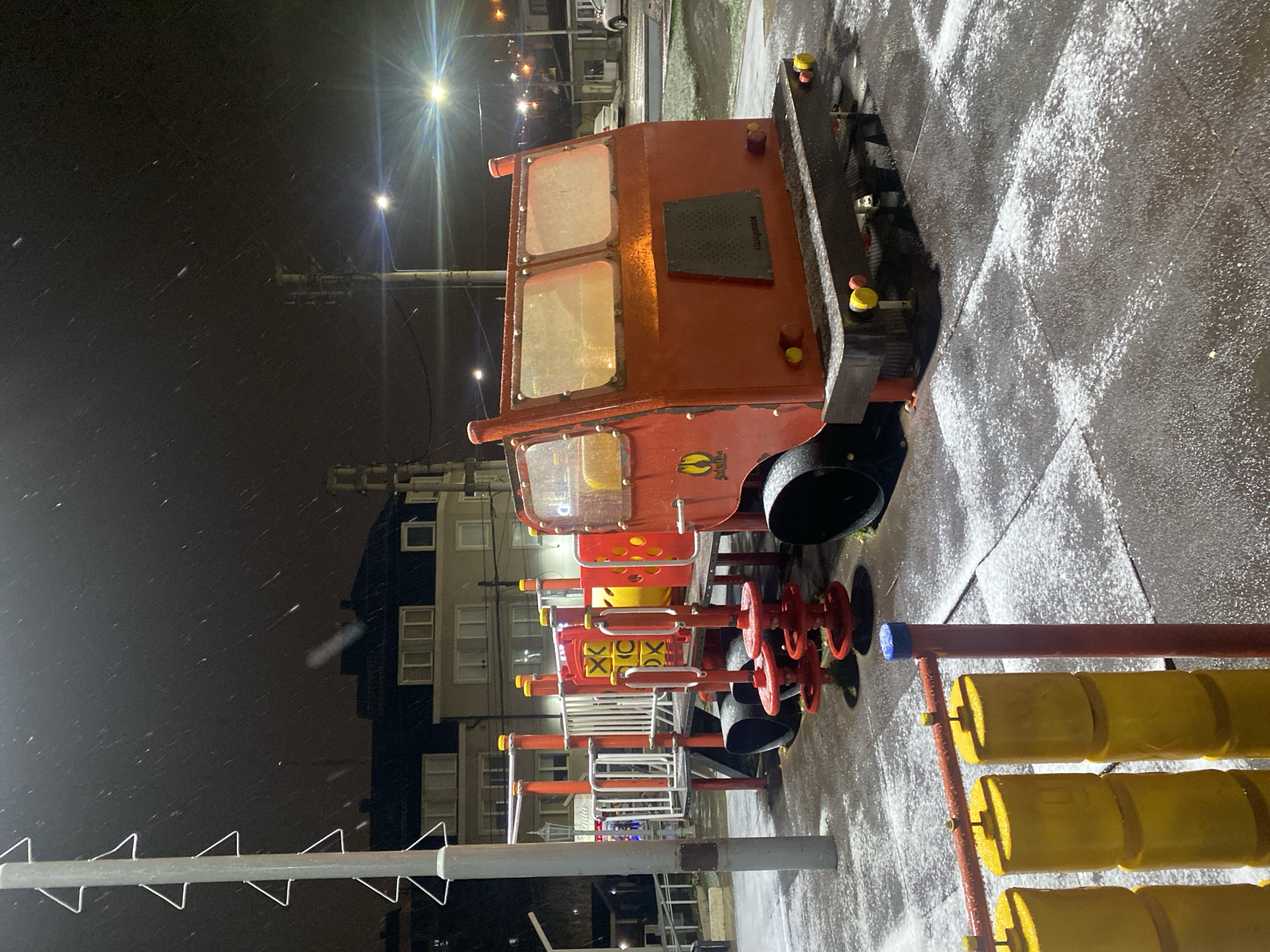 At night, in Ushuaia, Argentina, a snow-covered playground is illuminated by streetlights. The foreground features a multi-colored play structure, including a red train engine with a cabin, wheels, and various interactive elements like yellow cylinders and a tic-tac-toe board. Light snow is falling, visible in the dark sky and accumulating on the ground and play equipment. In the background, buildings and streetlights are faintly visible.