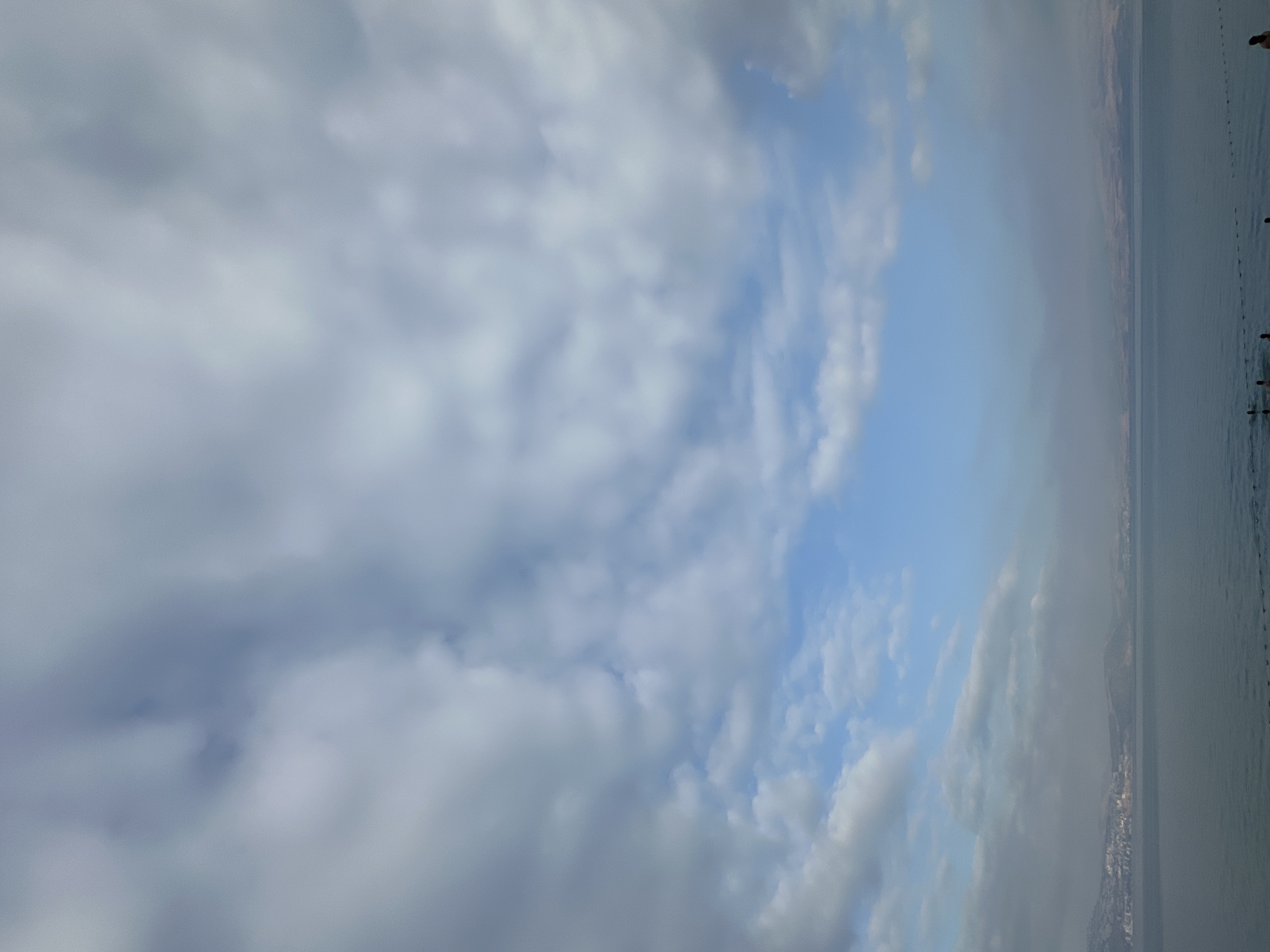 The image depicts a serene seascape with a wide expanse of calm water under a dramatic sky filled with various cloud formations. The clouds range from dense, grey masses to lighter, scattered patches, with glimpses of clear blue sky peeking through. In the distance, a hazy coastline is visible, characterized by low-lying hills and what appears to be a city or settlement along the shore. The water is a muted blue-grey, reflecting the overcast sky. Along the edge of the water in the foreground, several figures are present, some appearing to be in the water, suggesting a recreational activity. The overall atmosphere is tranquil and somewhat moody, with the vastness of the sky and sea dominating the composition. The presence of the distant coastline, coupled with the water, strongly suggests this scene is the Sea of Galilee, with the city of Tiberias being a prominent location on its western shore.