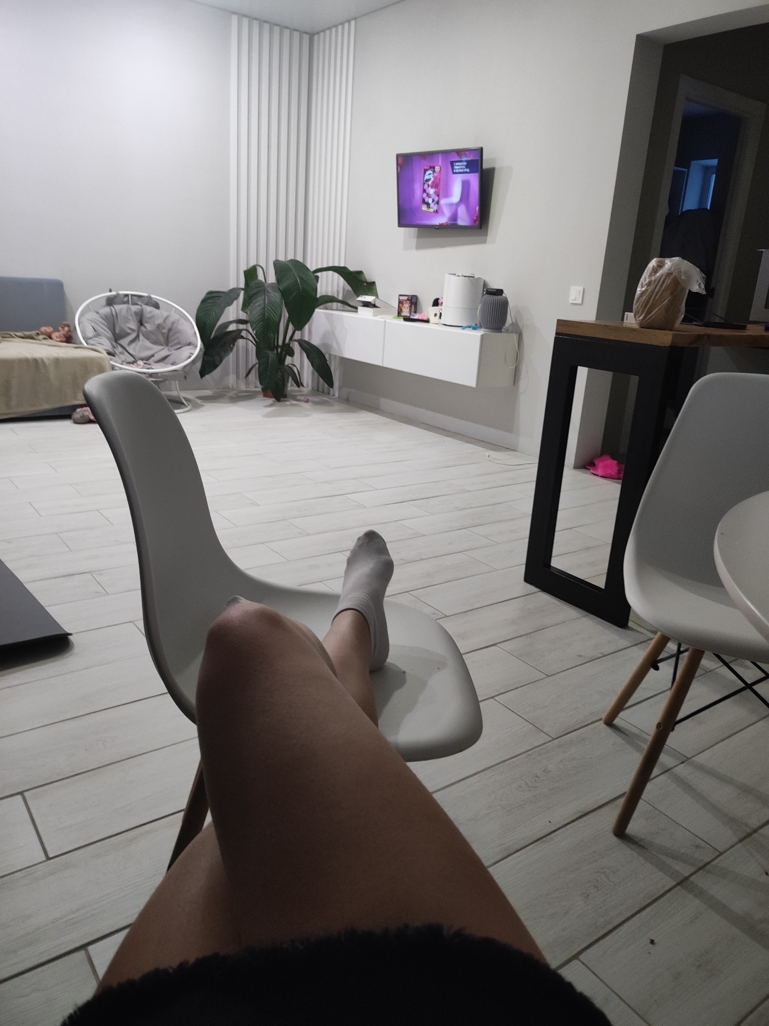 A person is seated in a white chair in an indoor space. The person's legs are crossed, and they are wearing a sock on their left foot. In the background, there is a living room setting with a sofa, a white cradle chair, a plant, and a white cabinet under a wall-mounted television. To the right, there is a black-framed table with a white chair next to it. The flooring appears to be light-colored tiles. There is no explicit information to determine the specific location in the city of Votkinsk, Russia.