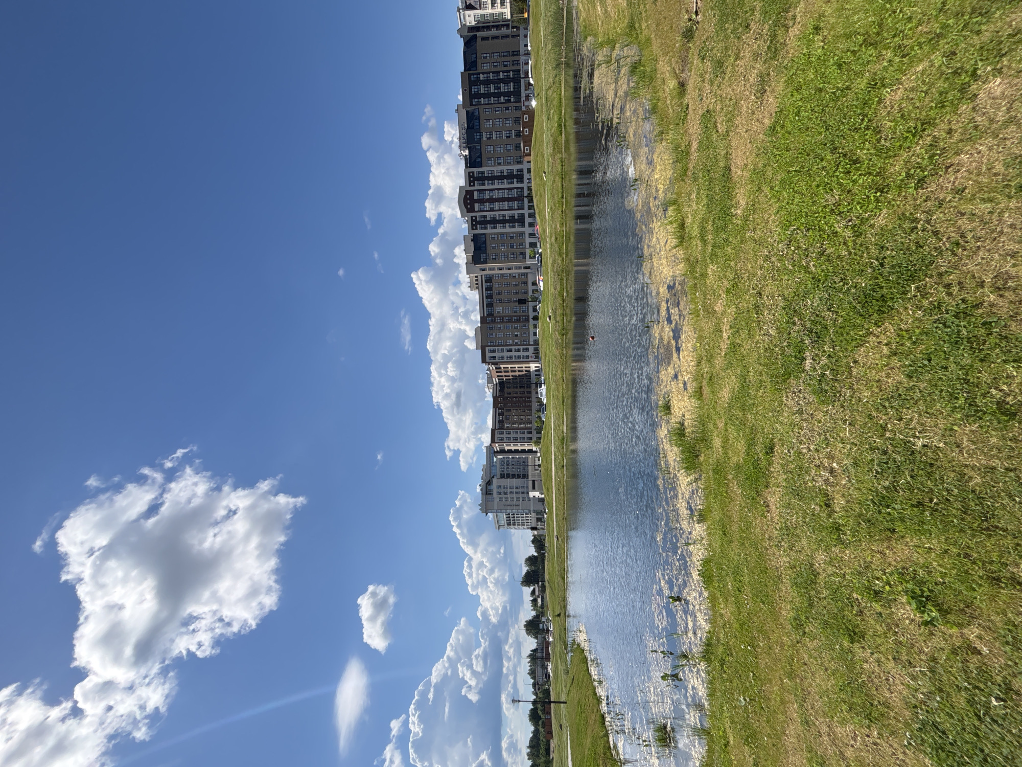 The image depicts a scene near a body of water, possibly a pond or lake, with buildings visible in the background. The sky is blue with scattered white clouds. Green and tan grass is on one side of the water. The buildings in the background appear to be multi-story residential or commercial structures. Based on the provided location, this is in Varaksino, Russia.