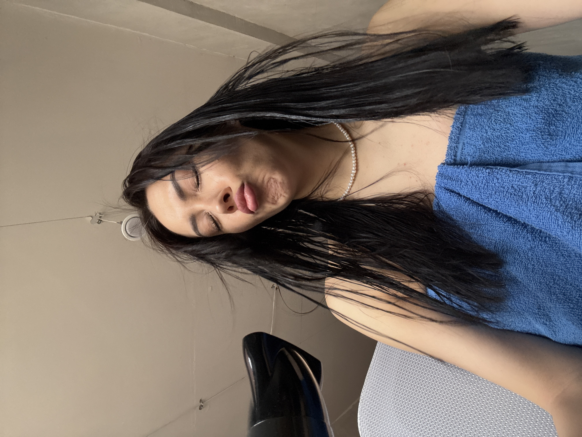 The image shows a woman with long, dark hair wearing a pearl necklace and a blue towel. Her eyes are closed, and her lips are pursed. A black hair dryer is visible in the lower left corner of the image. The background appears to be a neutral-colored wall and ceiling. The location in Votkinsk, Russia, is not discernible from the image.