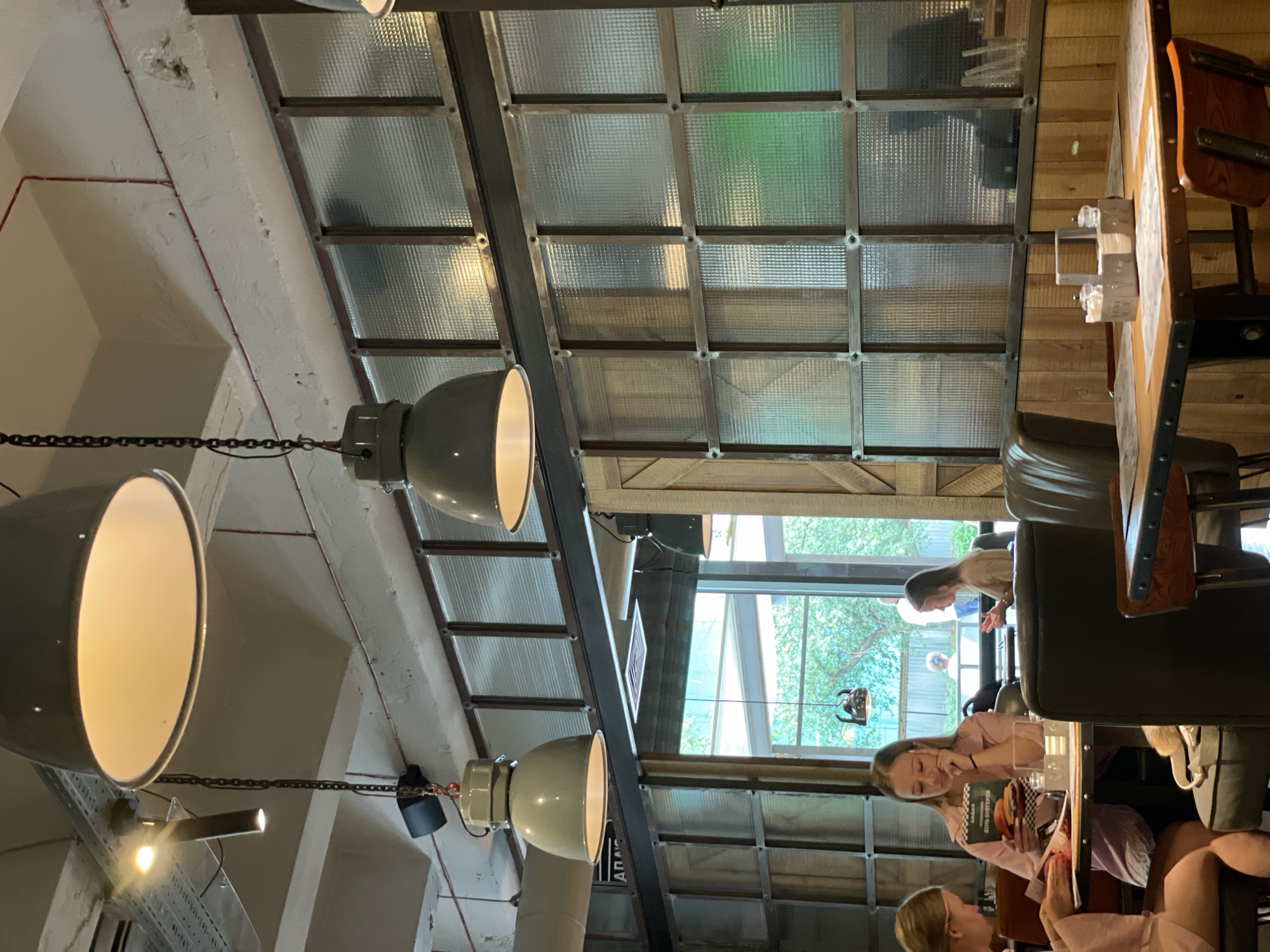 The image depicts the interior of a restaurant with an industrial aesthetic. Several large, pendant light fixtures hang from the ceiling. The walls are a mix of exposed concrete, dark wood paneling, and large windows with patterned glass divided by metal frames. In the foreground, a table is set with placemats, cutlery, and a napkin holder. Three young women are seated at tables, engaged in conversation or looking at menus. One woman on the left is wearing a light pink top and is looking at a menu. Another woman in the center is wearing a similar pink top and holding a menu, appearing to read it. A third woman in the background is wearing a beige top and looking towards another table. The overall lighting is warm, suggesting an evening or indoor setting. The scene is likely a restaurant in Krasnoyarsk, Russia, based on the provided context, though this cannot be visually confirmed from the image alone.
