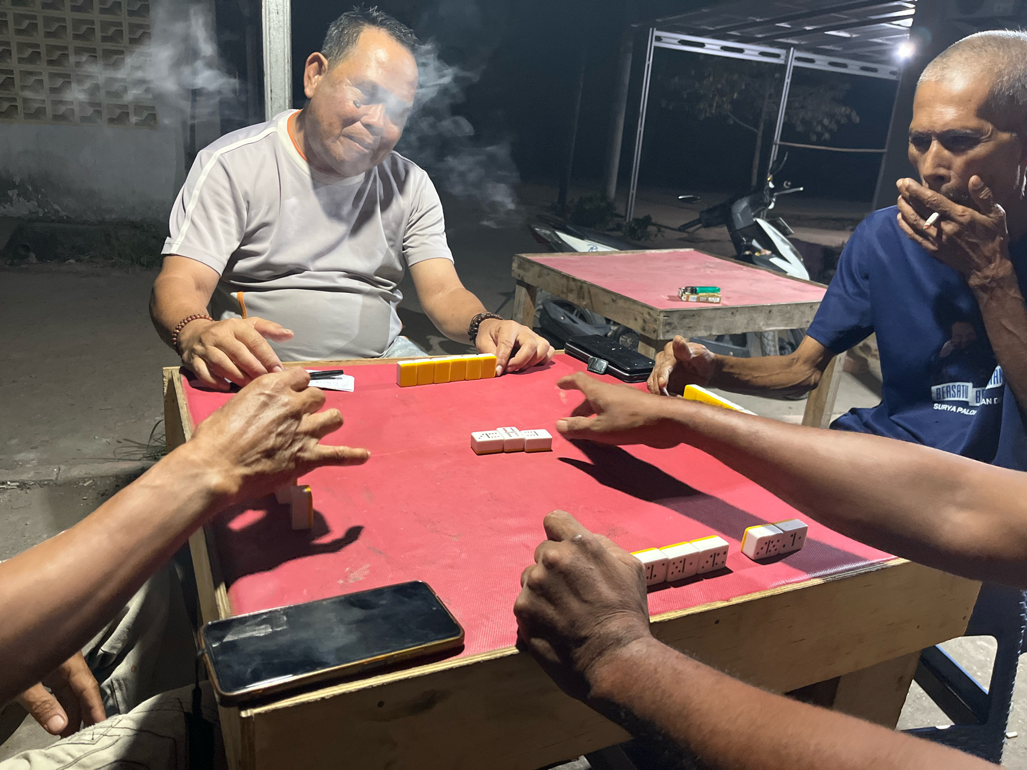 An outdoor scene at night in Talang Kelapa, Indonesia, depicts four men playing a game of dominoes around a red-covered table. Two of the men are smoking. The table has several domino tiles arranged on it, along with a mobile phone. In the background, there are structures, a motorcycle, and foliage. The lighting is dim, suggesting artificial illumination.