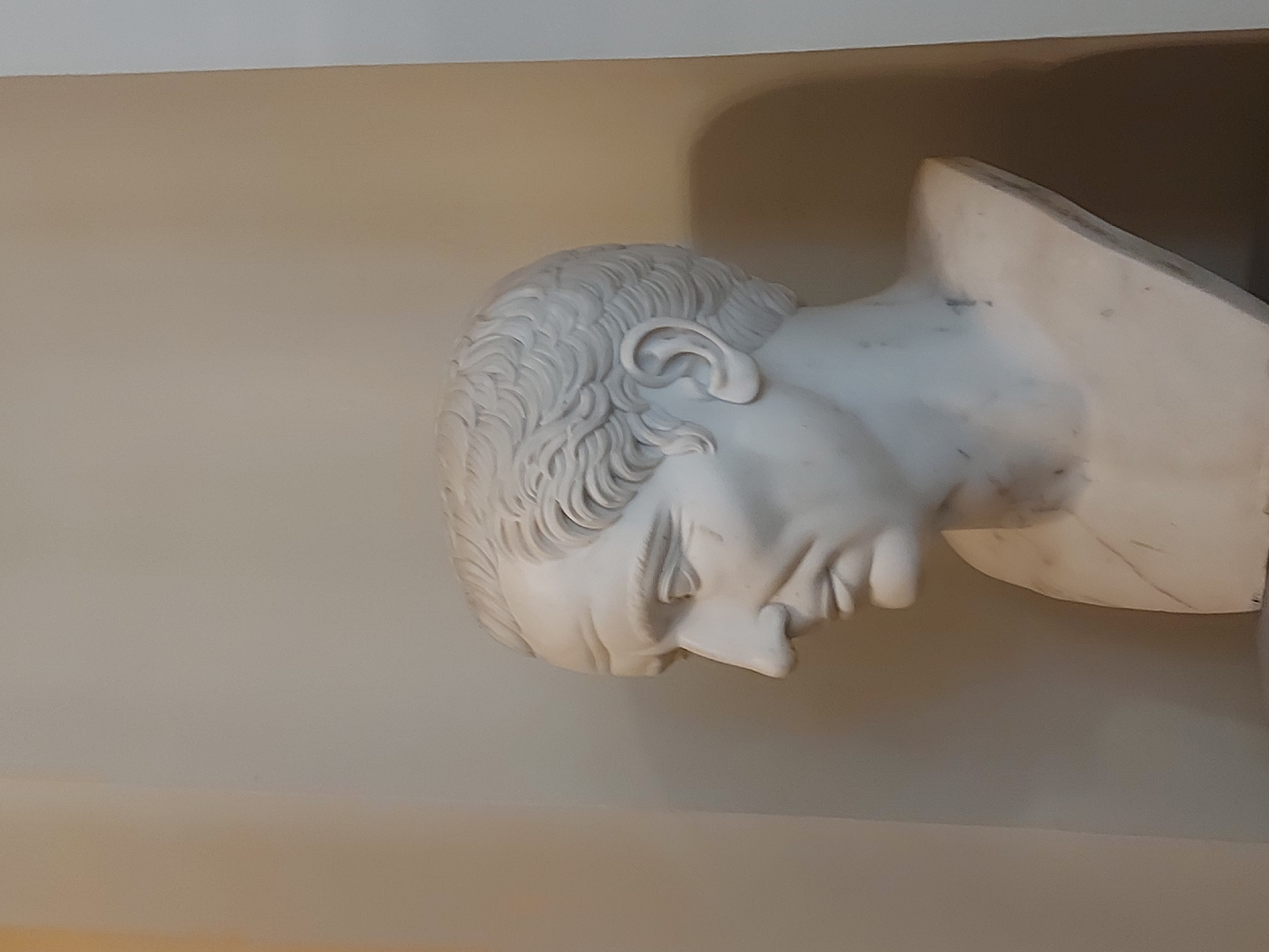 A close-up shot displays a white marble bust of a man with a sharply defined nose, curly hair, and an ear visible on the left side of his head. The bust is set against a light beige or off-white wall. The angle of the shot positions the bust slightly askew, as if it is mounted on a wall. The image was taken at The Yusupov Palace on the Moika, St Petersburg, Russia.