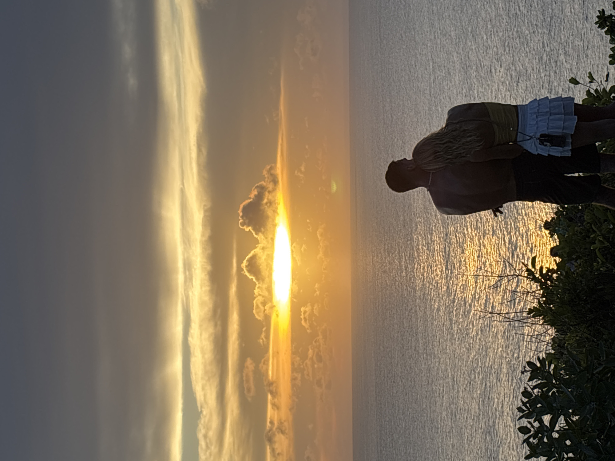 A couple stands on the edge of a cliff overlooking the ocean, silhouetted against a bright sunset. The man is shirtless, wearing dark pants, while the woman has blond hair and wears a green top and white ruffled skirt. They are embracing. The sun is low on the horizon, shining brightly through the clouds and reflecting on the water. Vegetation is visible on the cliffside. The location is Uluwatu, Indonesia.