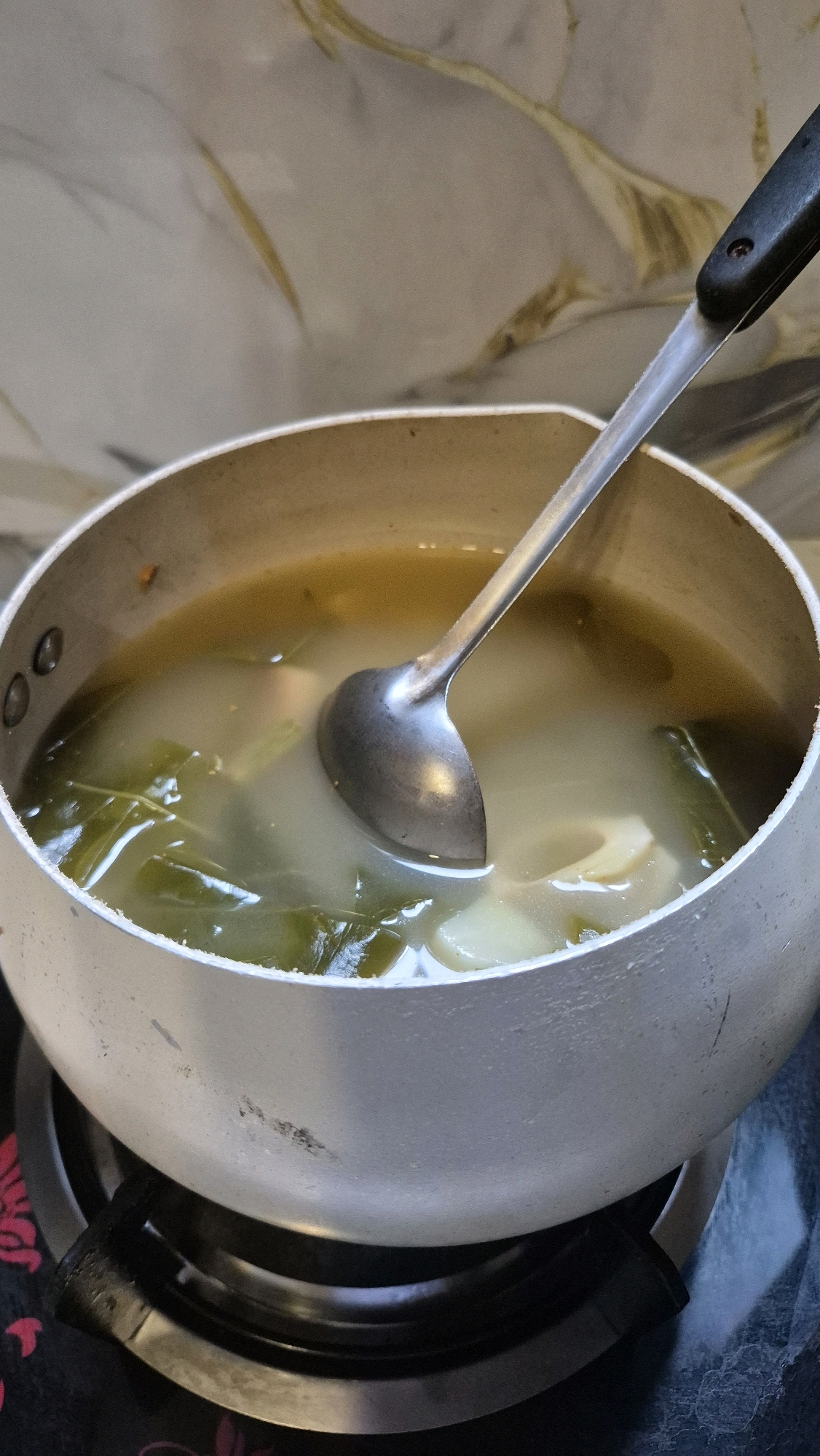 An overhead medium shot captures a stainless steel pot filled with a light-colored broth containing green leafy vegetables and sliced white ingredients, possibly bamboo shoots, situated on a black gas stove burner. A metal ladle with a black handle is partially submerged in the broth, its curved bowl resting at the bottom of the pot. The pot itself shows signs of use, with some scuff marks on its exterior. The background features a marbled countertop. The scene is consistent with a domestic kitchen setting in South Tangerang, Indonesia.