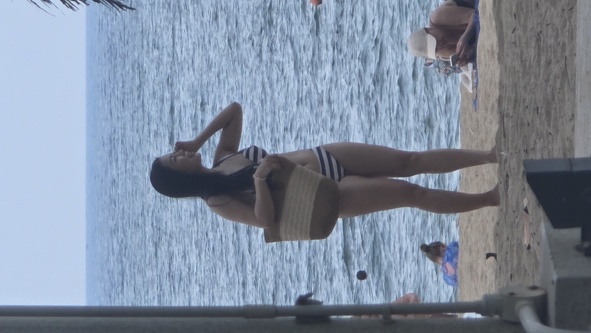 The image shows a woman standing on a beach in Nha Trang, Vietnam. She is wearing a striped bikini and holding a straw bag. Her left hand is near her head. The ocean is behind her. In the distance, there is a man in a white baseball cap looking at his phone while lying on the beach. In the foreground, there are architectural elements of a nearby structure. Another figure is visible further down the beach.