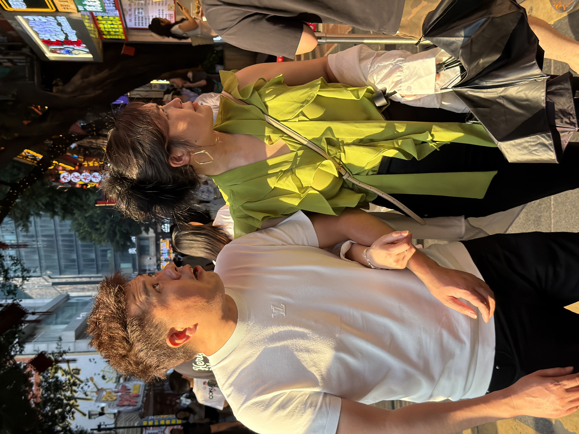 The image shows two people, a man and a woman, standing on a street at what appears to be dusk or evening. The man is in the foreground, wearing a white t-shirt with a small "LV" logo on it and black pants. He has his mouth open and is looking upwards and to the left. The woman is behind him and to his left, wearing a green ruffled top and black pants, also looking upwards and to the left. A light-colored purse is slung across her body. A black umbrella is partially visible behind the woman. In the background, there are illuminated signs for businesses, trees, and other people walking on the street. The lighting suggests it is nighttime or late evening, with artificial lights from the signs and streetlights illuminating the scene. The overall atmosphere seems to be that of a busy urban street.