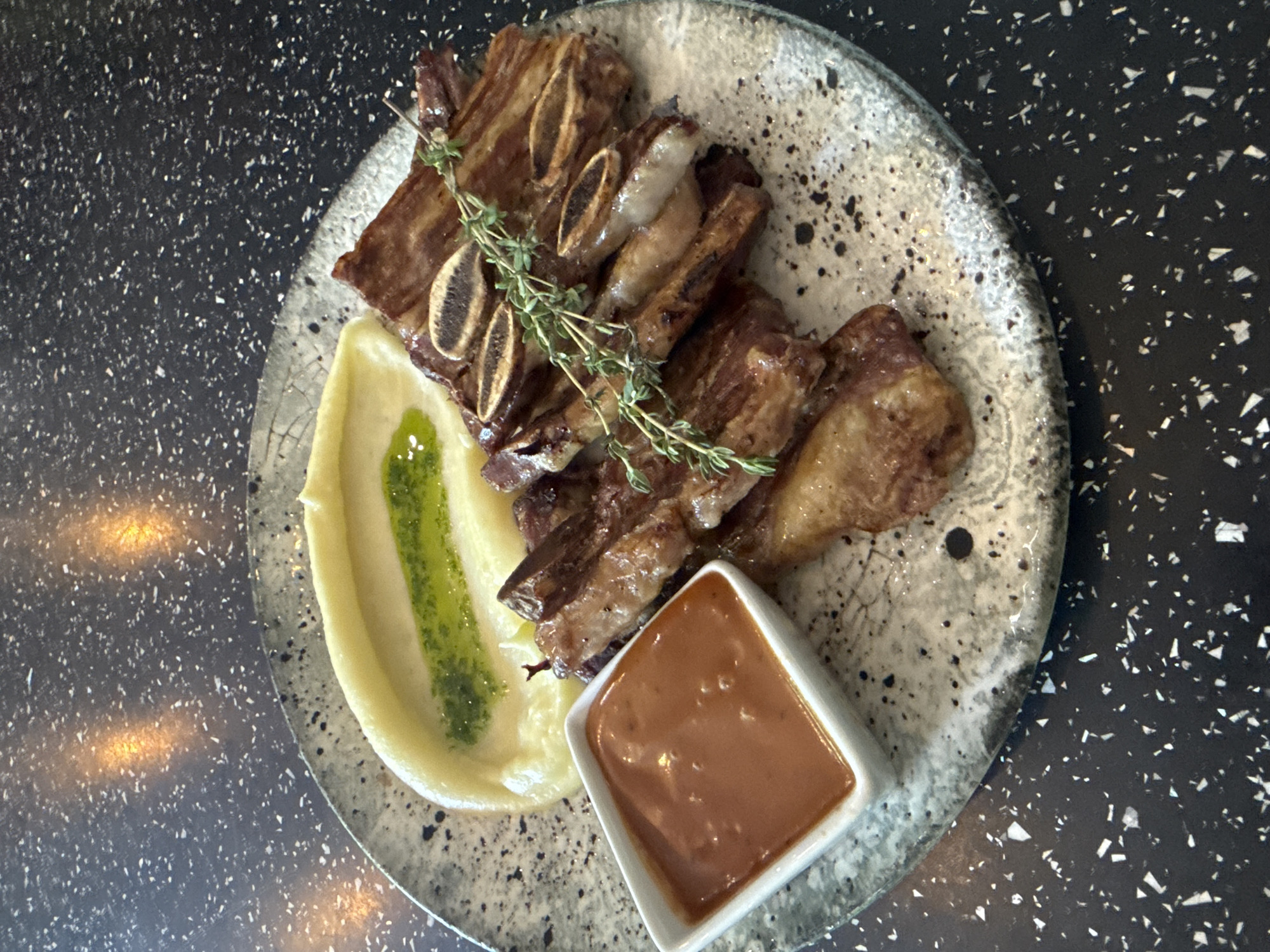 A close-up, overhead shot shows a serving of short ribs with mashed potatoes and a side of sauce, plated on a speckled gray dish. The short ribs, a deep brown with visible marbling and bone sections, are arranged in a fan-like pattern. A sprig of fresh thyme garnishes the ribs. To the left of the ribs is a dollop of smooth, pale yellow mashed potatoes, topped with a thin drizzle of vibrant green sauce. In the lower left corner, a small white square ramekin holds a reddish-brown sauce. The plate rests on a black speckled countertop, with indistinct orange and yellow reflections visible in the upper portion of the background, possibly from overhead lighting. The scene is static and appears to be an indoor setting, likely a restaurant. There are no people or animals present in the image. The culinary presentation suggests a focus on the food itself. The location is not specified in the image but is presumed to be in Varna, Bulgaria, as per the prompt.