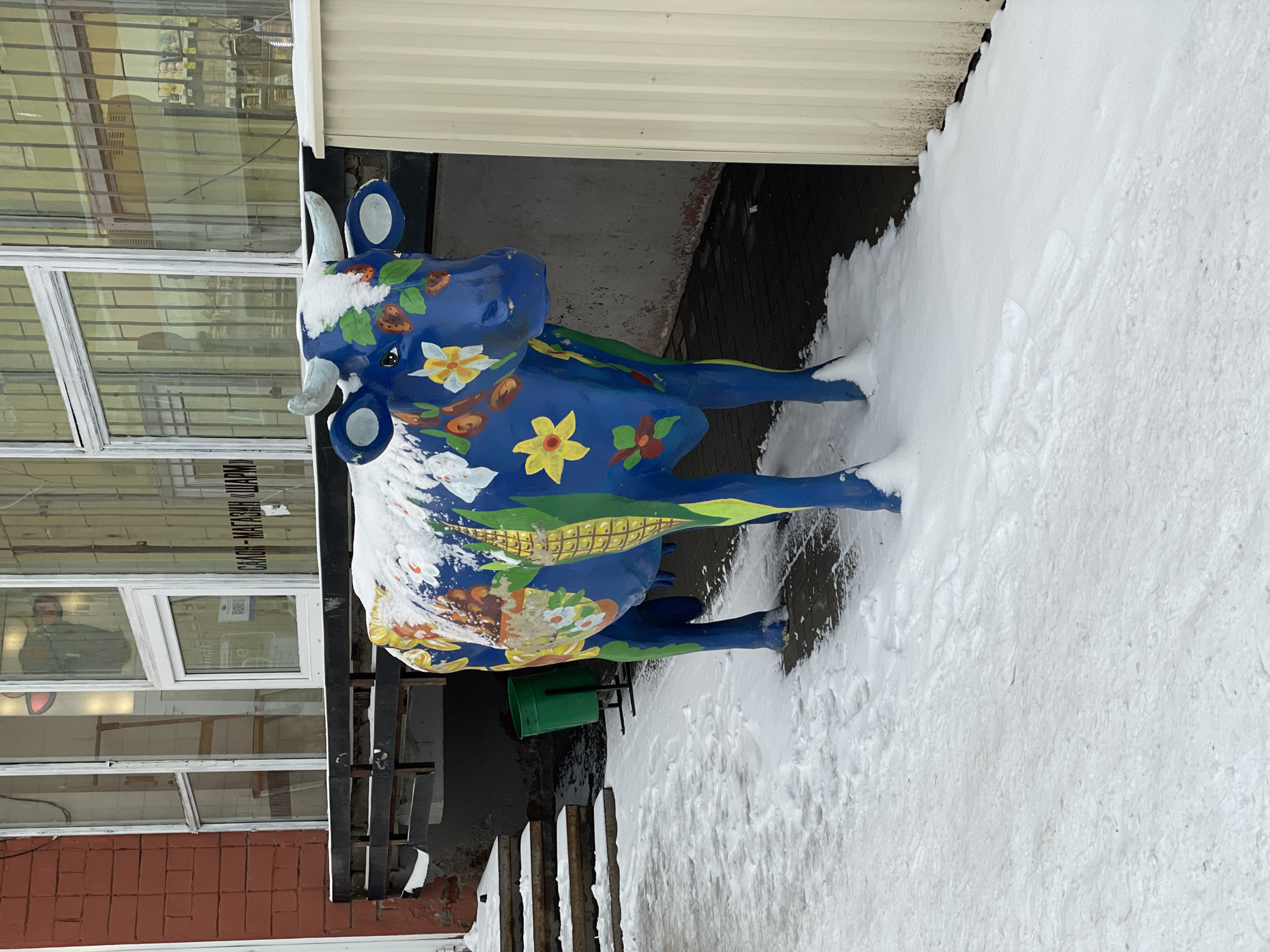 The image captures a blue and multi-colored painted cow statue standing near a building, partially covered in snow. The statue is adorned with floral patterns, fruits, and other colorful decorations. The building appears to be a commercial establishment, with windows and possibly signage visible on the left side. There is a substantial amount of snow on the ground around the statue, indicating winter conditions. The location is identified as Izhevsk, Russia, based on text visible on the building. There is a glimpse of a green bucket next to the statue. The image is taken from a medium distance, providing a clear view of the statue and its immediate surroundings.