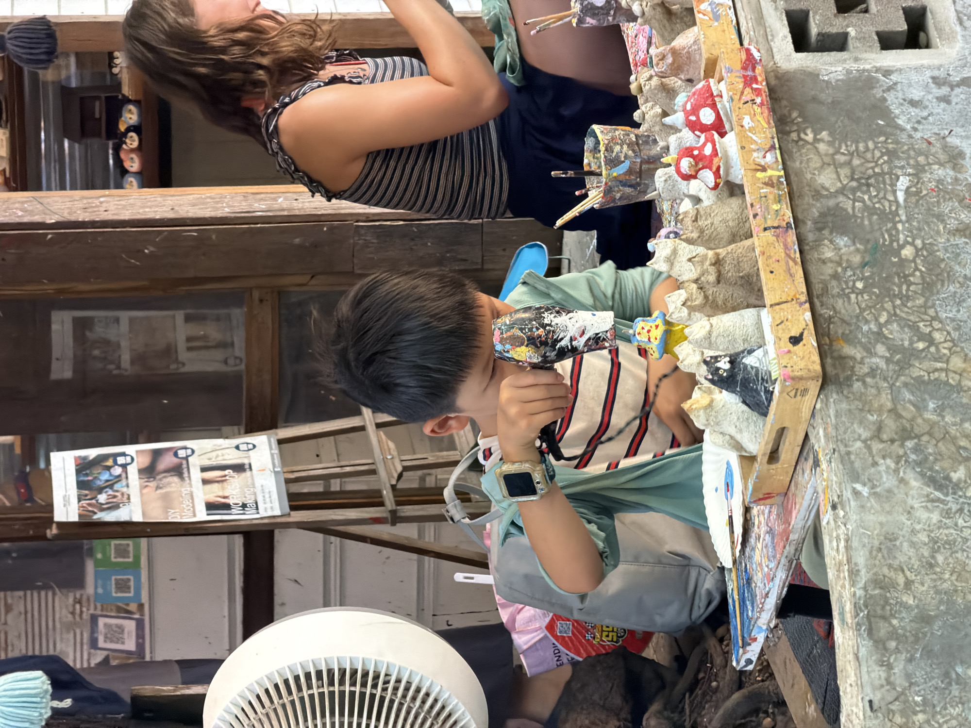 In Ban Sai Kham, Thailand, a young boy with dark hair, wearing a striped t-shirt and a light green overshirt, is focused on painting small ceramic figures on a wooden table. He has a smartwatch on his left wrist. To his right, a woman with long brown hair, wearing a striped tank top, is also engaged in painting. The table is cluttered with various art supplies, including paintbrushes, paint pots, and numerous unpainted and painted ceramic figures, many of which resemble mushrooms and animals. In the background, a large white fan is visible, along with posters advertising "DIY Modeling" and "Workshop Making," suggesting the location is an art studio or workshop. The scene appears to be outdoors, with natural light illuminating the area.