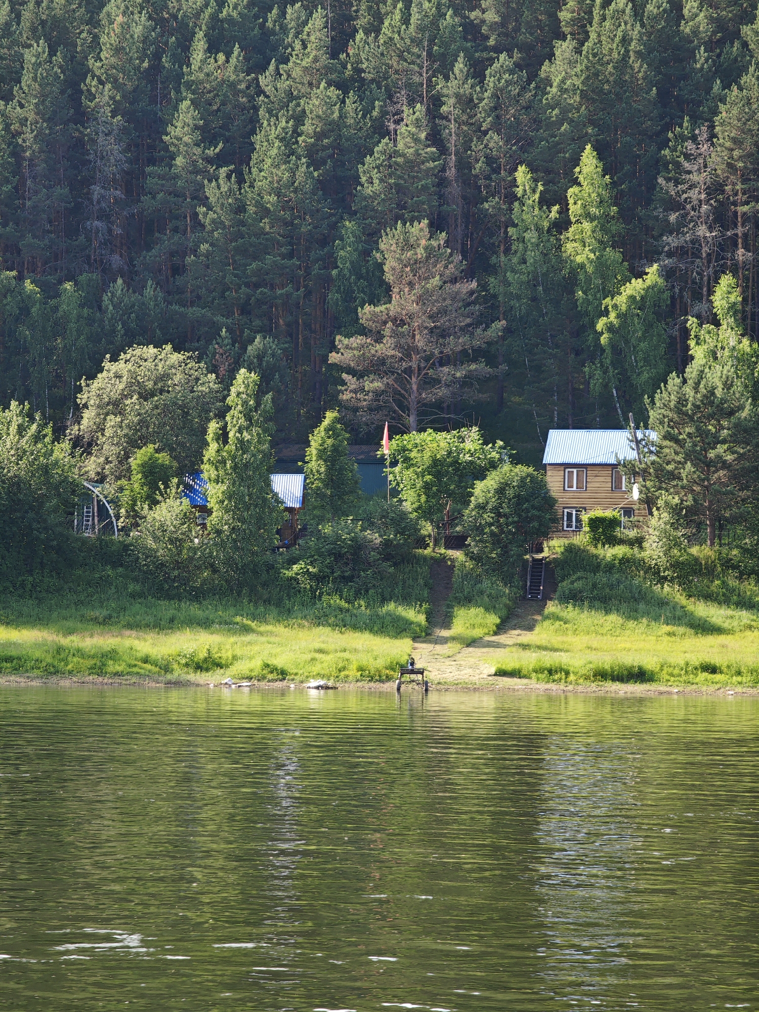 The image depicts a serene lakeside scene with lush green vegetation and a dense forest in the background. In the foreground, the calm water of a lake reflects the surrounding greenery and the bright sky. A narrow strip of grassy shore separates the water from a gentle slope leading up to a cluster of buildings nestled amidst trees. Two wooden houses with blue roofs are visible, one larger and two-storied, and another smaller, single-story structure. A pathway with wooden steps ascends the slope towards the houses. A red flag is planted near the smaller building. On the shore, a small, dark object, possibly a bench or a piece of equipment on wheels, sits near the water's edge. The overall impression is one of a peaceful, rural retreat in a natural setting, likely a dacha or a small settlement along the riverbank in Pristan', Russia.