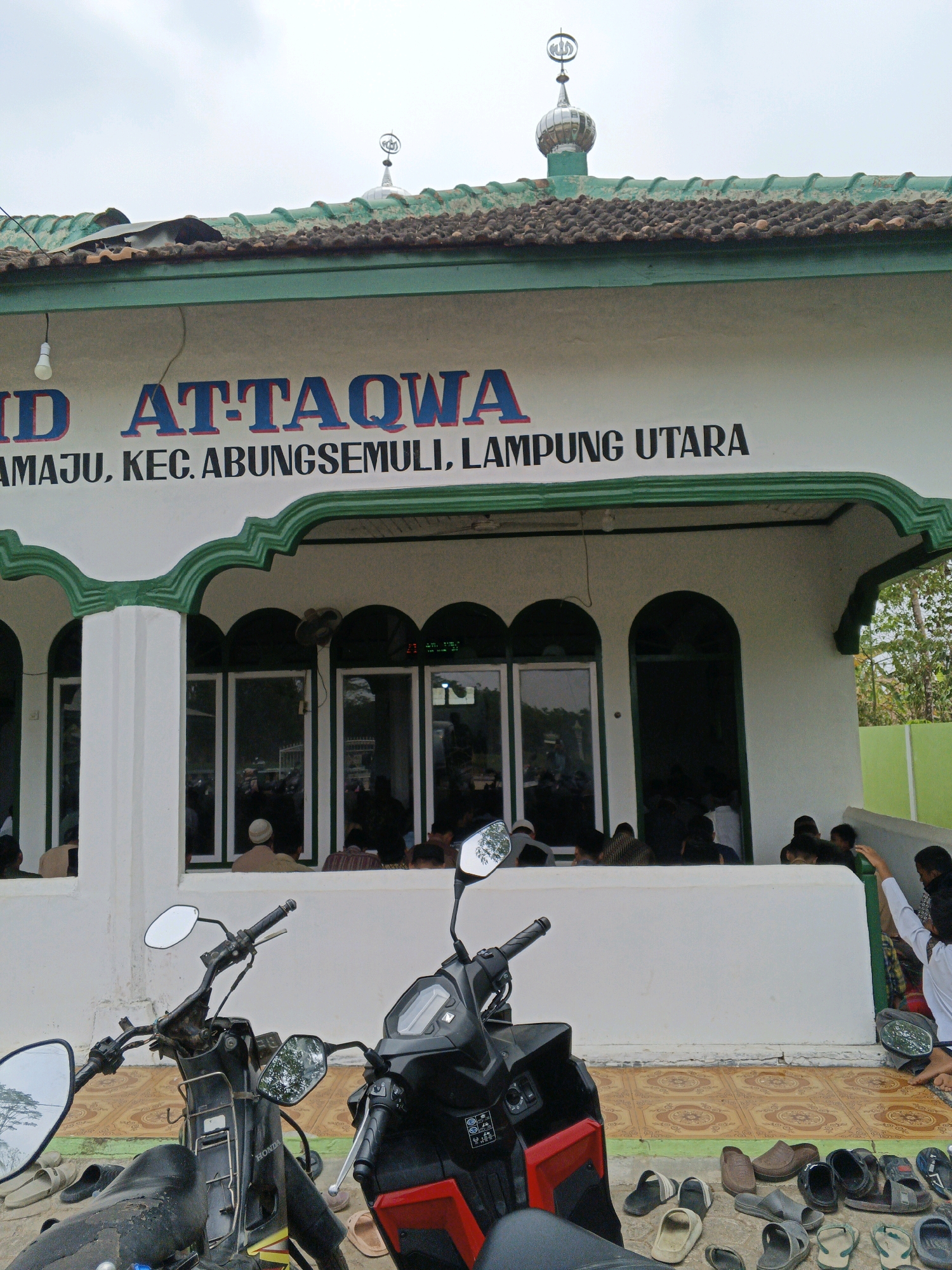 The image shows the exterior of a building identified by signage as "MASJID AT-TAQWA". Below this, text reads "AMAJU, KEC. ABUNGSE MULI, LAMPUNG UTARA", indicating the location as Abungsemuli District, North Lampung Regency, Lampung Province, Indonesia. The building has a white facade with decorative green trim and a tiled roof. A small dome with Islamic calligraphy is visible on the roof. In front of the building, two motorcycles are parked. Several pairs of footwear are scattered on the ground in front of the building's entrance. Inside the building, visible through open doorways and windows, a number of people are gathered, appearing to be engaged in prayer or other religious activities. The ground in front of the building is paved with decorative tiles. The sky is overcast.