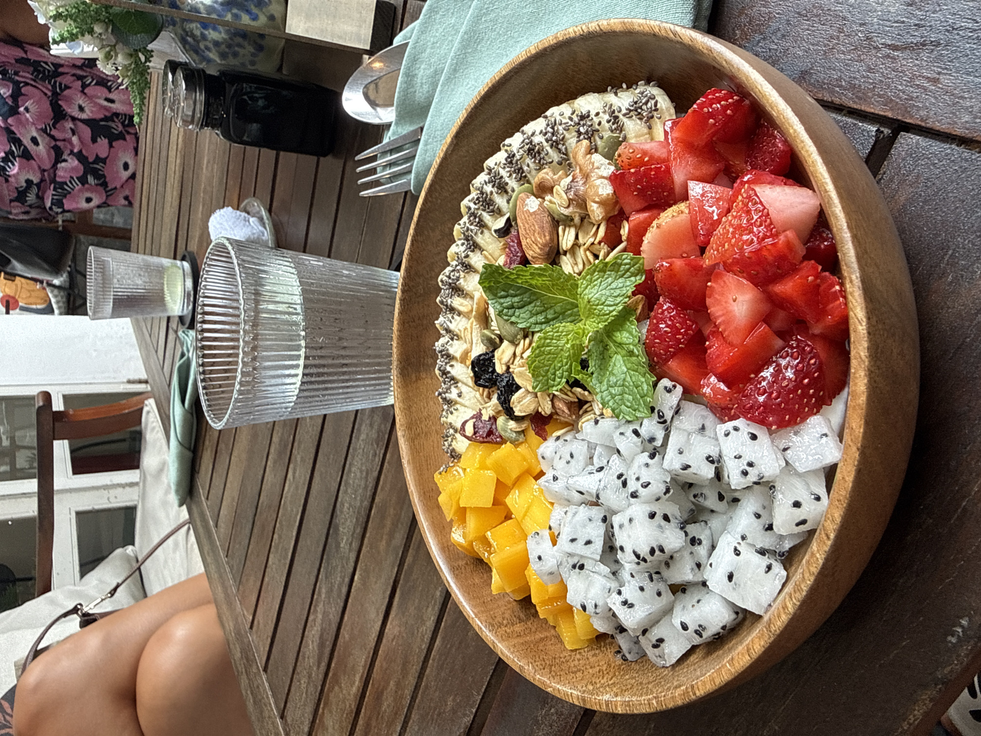 A high-angle shot shows a wooden table with a wooden bowl filled with fruit, granola, and a mint garnish, alongside glasses and cutlery. The bowl contains sliced strawberries, cubed dragon fruit, cubed mango, banana slices with chia seeds, and granola with nuts and dried fruit. The table, made of wooden slats, also holds two cylindrical, vertically ribbed glasses, a black jar, and a fork and spoon. A light green cloth napkin is partially visible beneath the cutlery. To the left, a person's knees and handbag are visible near a white cushioned chair and a window with white trim. The specific location within Đà Nẵng, Vietnam, cannot be determined from the image.