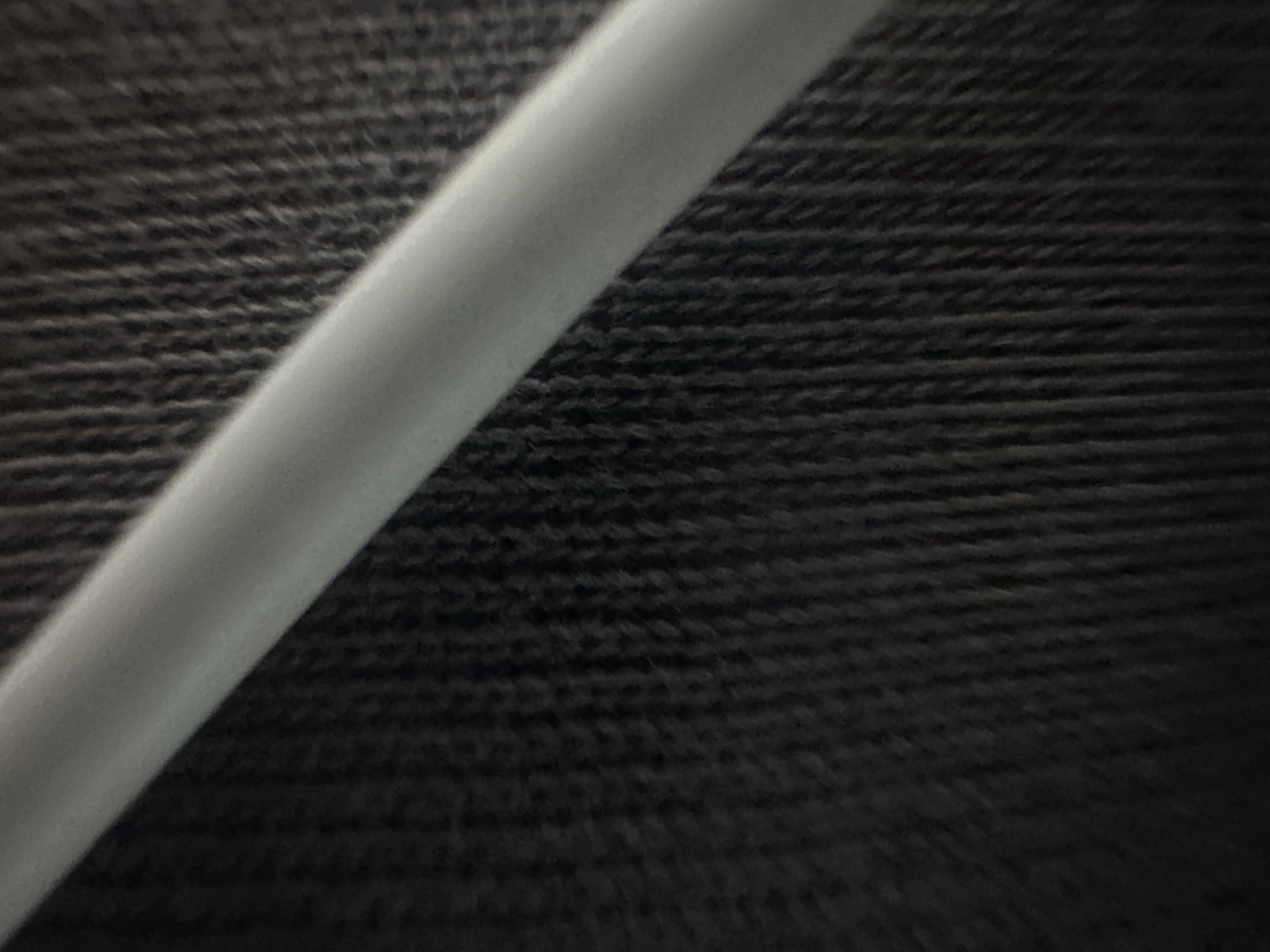 The media file is a close-up, macro photograph of a dark, textured fabric, likely denim, with a light gray, cylindrical object diagonally crossing the frame from the bottom left to the upper right. The fabric has a distinct weave pattern visible, characterized by horizontal lines of darker thread interspersed with lighter thread, creating a ribbed effect. The light gray object appears smooth and slightly curved, with some subtle shading indicating it is three-dimensional. The lighting suggests a soft, diffused light source, possibly from above and to the left, casting gentle shadows on the fabric. The focus is sharpest on the left side of the gray object and the fabric immediately around it, with a slight blur towards the right side of the frame, creating a shallow depth of field. There are no people, discernible scenes, or explicit location cues beyond the provided context. The image does not depict any activity or event, nor does it contain any visible text. The overall impression is abstract and textural, focusing on the interplay of light, shadow, and surface detail.