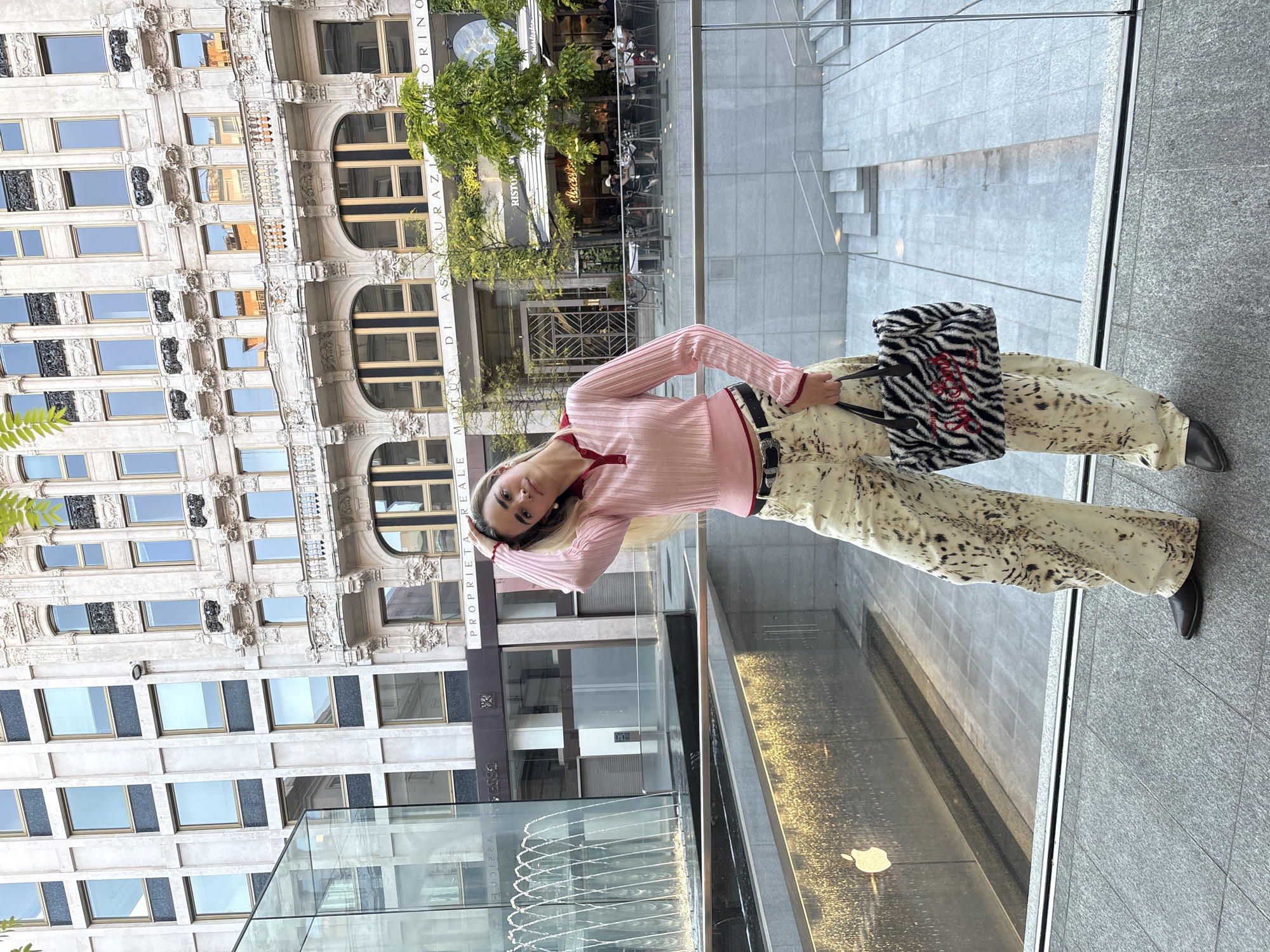A young woman is captured in a full-body shot, posing in an urban setting that blends historic and modern architecture in Milan, Italy. She has blonde hair and is dressed in a light pink, long-sleeved ribbed top with red trim, cream-colored wide-leg trousers featuring a dark animal print pattern, a dark belt with a prominent silver buckle, and dark pointed-toe shoes. She holds a black and white zebra-print tote bag with a red design, possibly 'LOVE' or a heart, on its side. Her right arm is raised with her hand near her head, and she looks directly at the camera.
Behind the woman, across what appears to be a street or open plaza, stands a grand, multi-story historic building. This structure boasts an ornate, light-colored stone facade with numerous rectangular windows, decorative balconies, and intricate architectural details typical of European cities. Text visible on the building includes "MUTUA DI ASSICURAZIONI," "ORINO," "RISTORAN..." (likely Ristorante), and "PROPRIETÀ REALI," which are Italian terms, supporting the Milanese context.
In the immediate foreground and to the woman's right, modern elements define the space. These include a clear glass barrier, grey stone flooring, and a distinctive water feature or fountain. The reflection of the Apple logo is clearly visible in the shimmering surface of the water feature. The scene is brightly illuminated, suggesting it is daytime with clear weather. The image conveys a fashionable and confident moment set against a dynamic architectural backdrop.