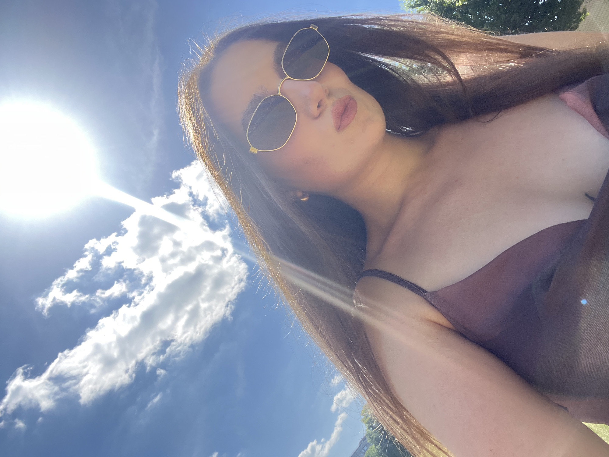In this close-up, low-angle shot, a young woman with long, brown hair is looking up at the sky. She is wearing sunglasses with gold frames and a dark-colored top. The sun is shining brightly in the upper left corner, casting a lens flare across the image. The sky is a vibrant blue with scattered white clouds. In the background, out of focus, trees and foliage are visible, suggesting an outdoor setting. The woman's facial expression is somewhat neutral, with her lips slightly parted. The lighting indicates it is daytime, likely a sunny afternoon. The image appears to be a selfie taken outdoors.