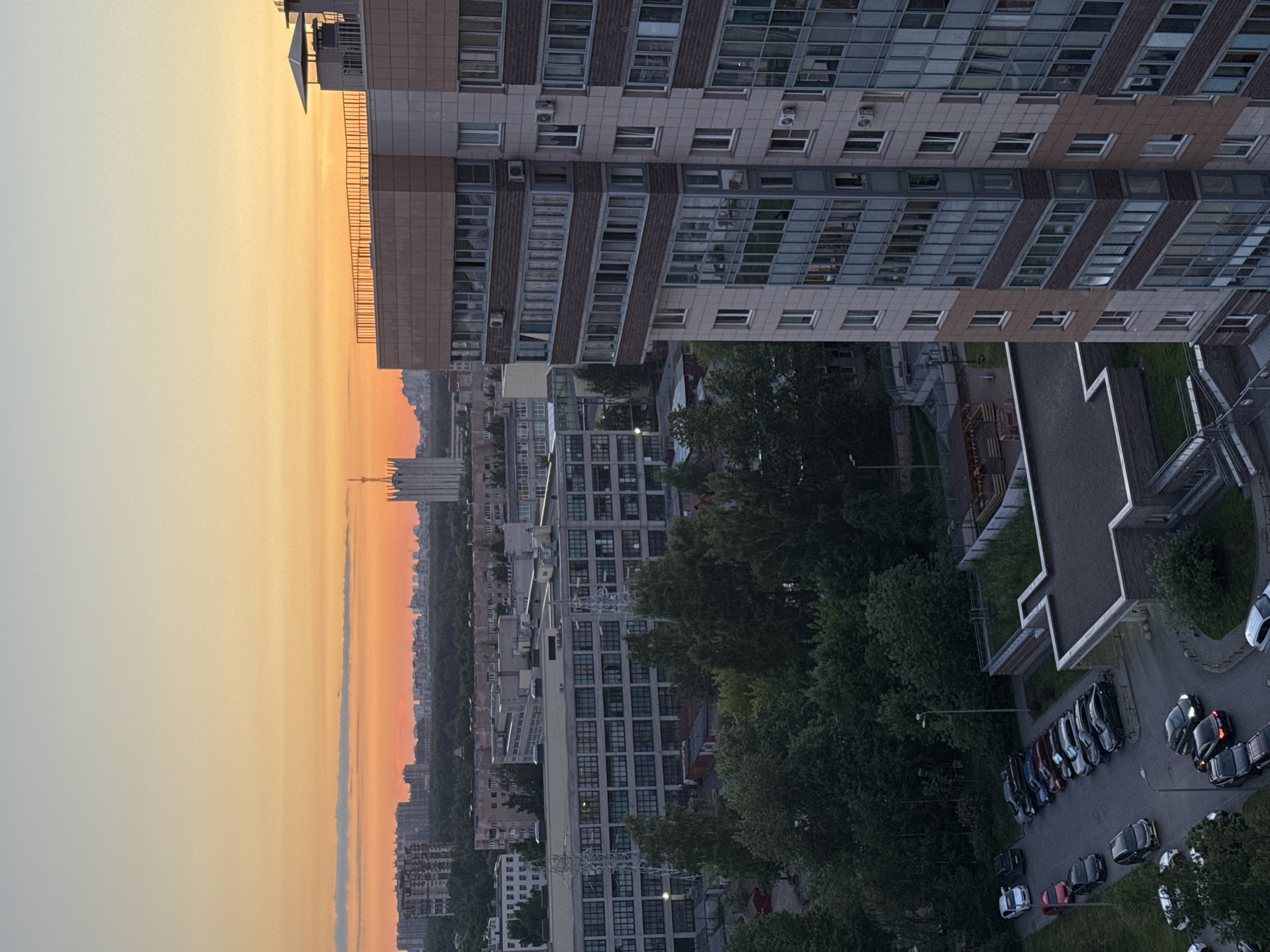 This image depicts an urban landscape in the city of Unknown, Russia, captured from an elevated viewpoint during sunset. The sky displays a clear gradient from a deep orange at the horizon, transitioning to yellow and pale blue overhead, with a few faint horizontal clouds present in the orange band.
In the foreground, occupying the right side, stands a modern, multi-story residential building with a facade comprising light beige panels, darker brown sections, and extensive glass windows and balconies/loggias. Numerous external air conditioning units are mounted on its exterior. The building features a flat roof with perimeter railings and visible ventilation structures.
Below and to the left of the prominent residential building, the midground contains various multi-story industrial or commercial buildings, predominantly grey with large window arrays, indicative of a mixed-use zone. Dense green trees and foliage are interspersed between these structures and along a treeline extending across the middle distance. Two distinct power line towers are visible within this area, flanking a central industrial building. In the lower-left foreground, multiple asphalt parking lots are populated with numerous parked cars of various models and colors. Green grass and scattered trees are present around these parking areas, which are illuminated by streetlights. A children's playground structure is partially visible behind a smaller building with a grey roof, located to the left of the main residential tower.
In the distant background, a dense city skyline is discernible, composed of numerous residential and commercial buildings. A distinctive, tall, light-colored building with a spire or antenna rises prominently above the other structures on the horizon. A broad expanse of dark green forest or parkland separates the midground development from the distant city. No individuals are explicitly visible within the scene.