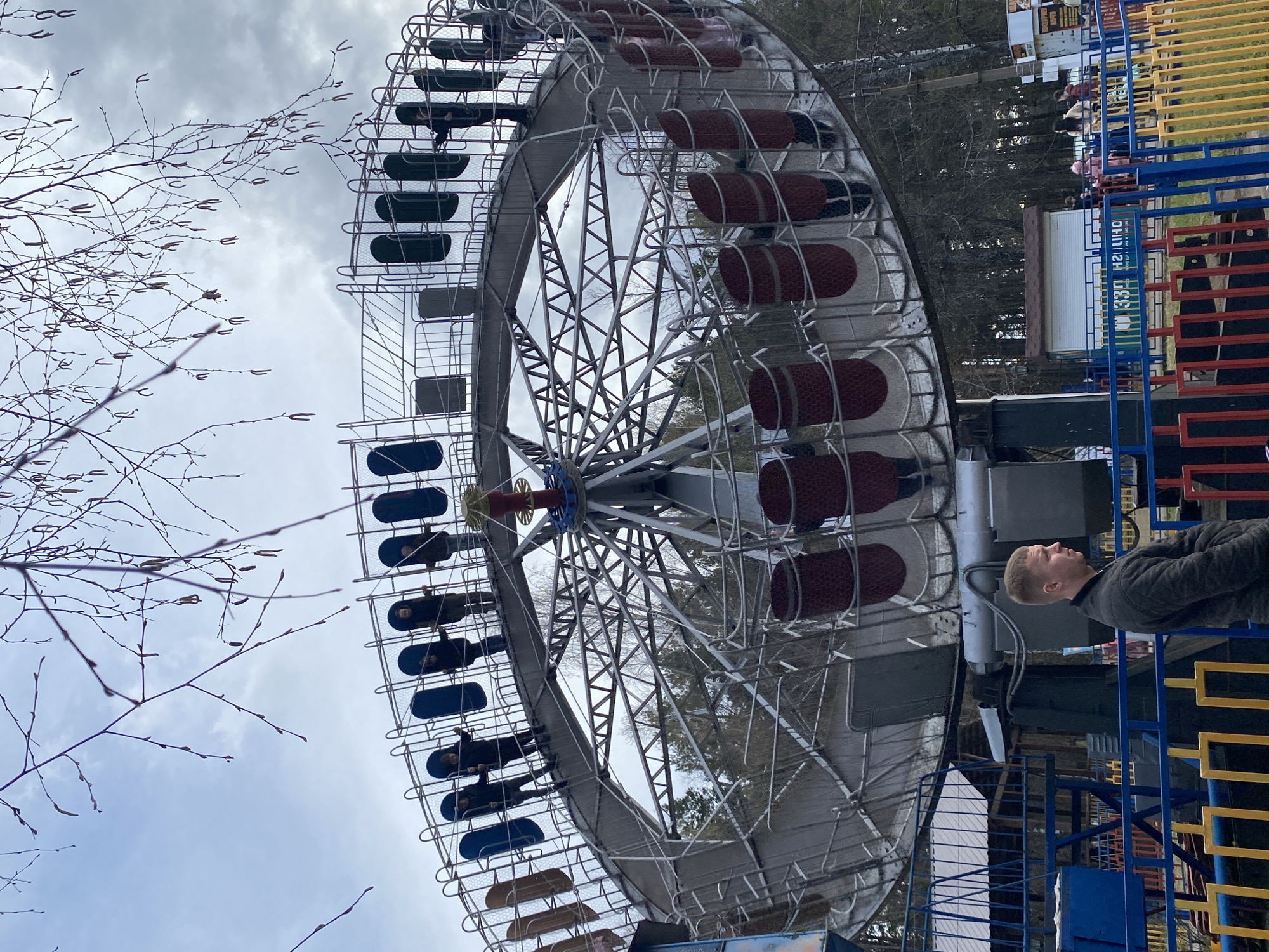 This image captures an amusement park scene under an overcast sky, likely in late winter or early spring due to the bare tree branches on the left. The central and dominant subject is a large, metallic amusement ride, appearing to be a variant of a Ferris wheel or a swinging pendulum ride, with numerous passenger compartments. The structure is predominantly silver-grey, with individual seats colored dark red, blue, and black. Several compartments contain people, some visibly children, suggesting the ride is either in operation or about to start.
In the foreground on the right, a man with short, light-colored hair, wearing a dark grey jacket, stands next to a vibrant barrier composed of blue, yellow, and red sections. He is looking towards the amusement ride. In the background, beyond the colorful fencing and the ride, trees with sparse branches and distant buildings are visible, including a small white structure with a brown roof.
A sign visible in the background, behind the man and the colorful barrier, displays Cyrillic text. Recognizable elements include "330", "H2", and "ЧАЙКОВСКИЙ" (Chaikovsky), confirming the location as Chaikovsky, Russia. The scene conveys a cool, daylight atmosphere in an outdoor recreational setting.
