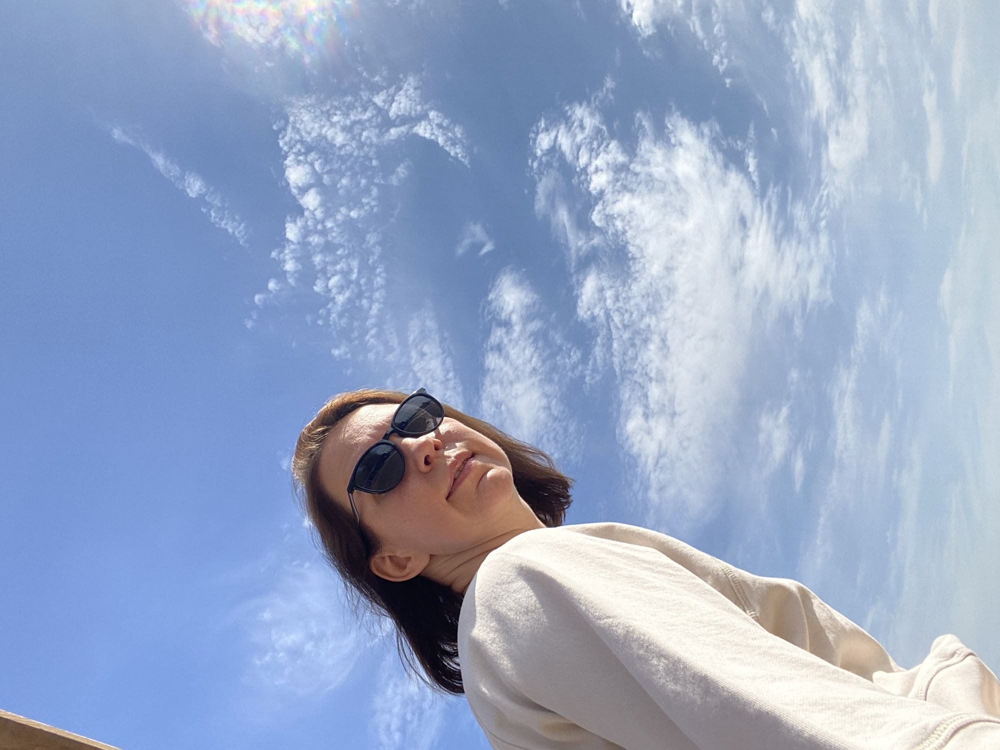The image is a low-angle selfie of a woman wearing sunglasses and a white sweatshirt. The background is a blue sky with scattered white clouds. The location of the image is not discernible from the provided information.