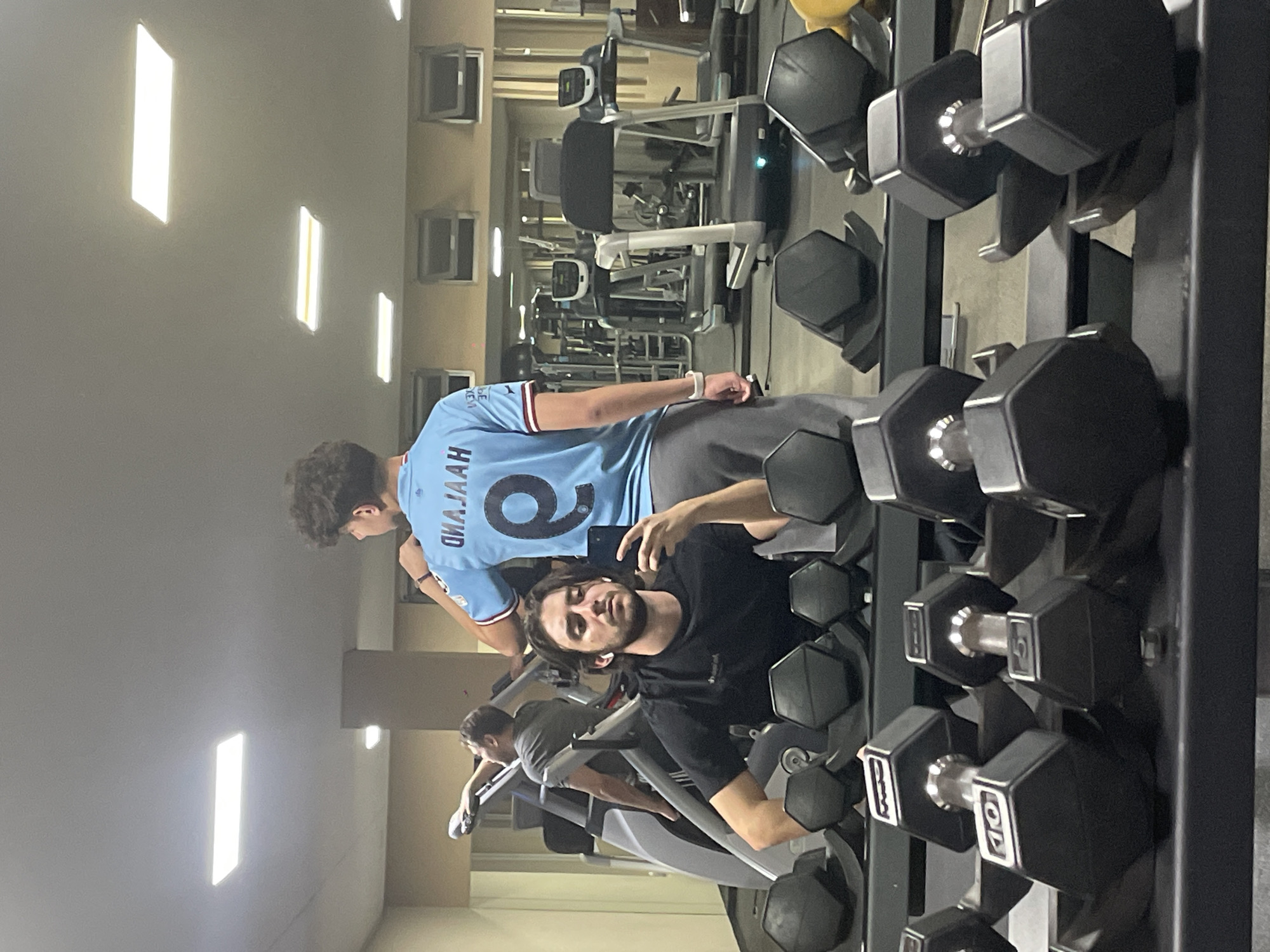 The image shows a gym setting with two people posing near a dumbbell rack. One person is wearing a light blue jersey with the number "9" and the name "HAALAND" on the back. The second person is wearing a black shirt and appears to be taking a selfie. In the background, there are various pieces of gym equipment, including treadmills and weight machines. The location is a gym, though the presence of windows suggests the building has residential or office space above it. There is no information to suggest that the gym is specifically located in the city of Huixquilucan de Degollado, Mexico.