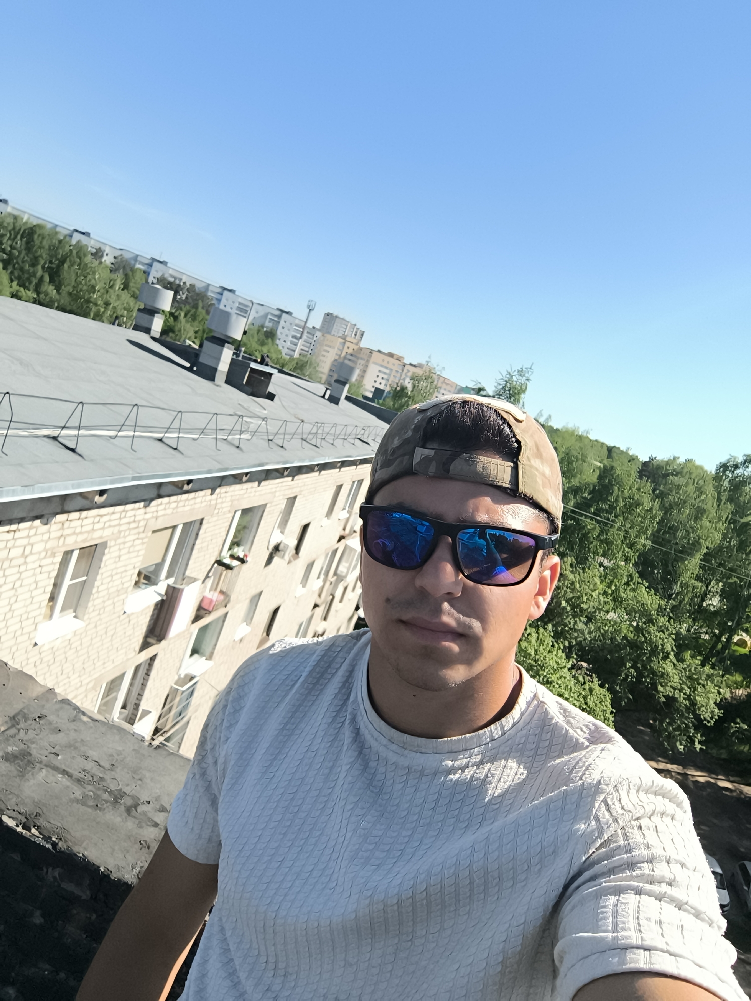 A person takes a selfie on a rooftop. The person is wearing sunglasses and a camo hat, and a patterned white t-shirt. Behind the person is the edge of a building, beyond which are trees and other buildings. The sky is blue and clear.