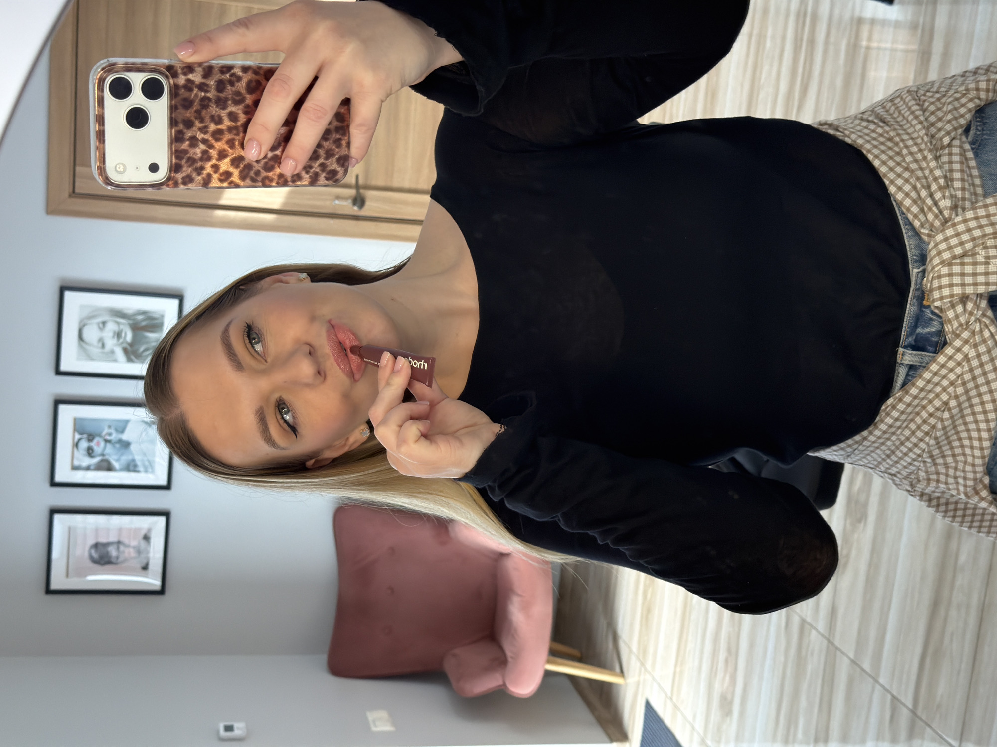 A woman is taking a selfie in what appears to be an indoor setting. She is holding a smartphone with a leopard print case in her right hand, and in her left hand, she is holding a lip gloss or lipstick tube, applying it to her lips. She is wearing a black long-sleeved shirt and a plaid shirt tied around her waist.
Behind her, there are framed pictures on the wall, and a pink chair is visible in the background. The flooring appears to be light-colored wood. The location cannot be determined based on the image, but the user requested it to be assumed as Riga, Latvia. Therefore, we can infer that this scene is purportedly taking place in Riga, Latvia.