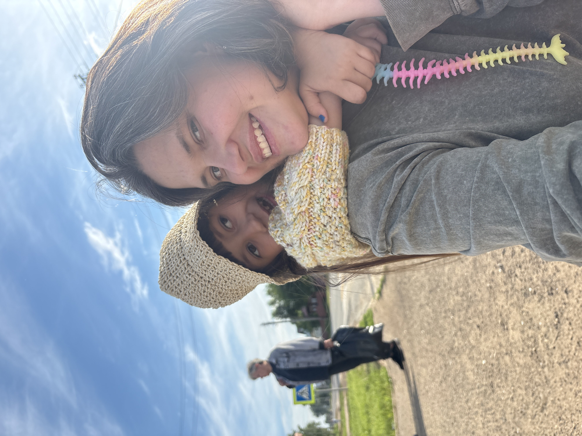 The image is a brightly lit, vertically oriented outdoor selfie featuring two people, likely an adult and a child, captured in Фоки, Russia. The adult, with dark hair showing some grey strands, is smiling broadly at the camera. They are wearing a grey long-sleeved top, and a distinctively textured, possibly hand-knitted, light yellow and multicolored cuff is visible on their arm. One fingernail is painted blue. The adult holds a rainbow-colored (blue, pink, and yellow) flexible toy, shaped like a fish skeleton or spine, near their shoulder.
Partially visible behind and below the adult is a child, wearing a light-colored, woven straw-like hat. The child looks towards the camera with a curious expression.
The background reveals an outdoor setting under a bright blue sky with scattered white clouds, suggesting sunny daytime weather. The lower part of the background shows an out-of-focus street scene, including a paved path or sidewalk, a grassy verge, and distant trees. A blurry figure of another person, an adult, is seen walking in the mid-ground. Near this figure, an indistinct blue and yellow road sign, possibly indicating a pedestrian crossing, is visible. The overall mood of the image appears cheerful, emphasized by the adult's smile and the clear, sunny conditions.