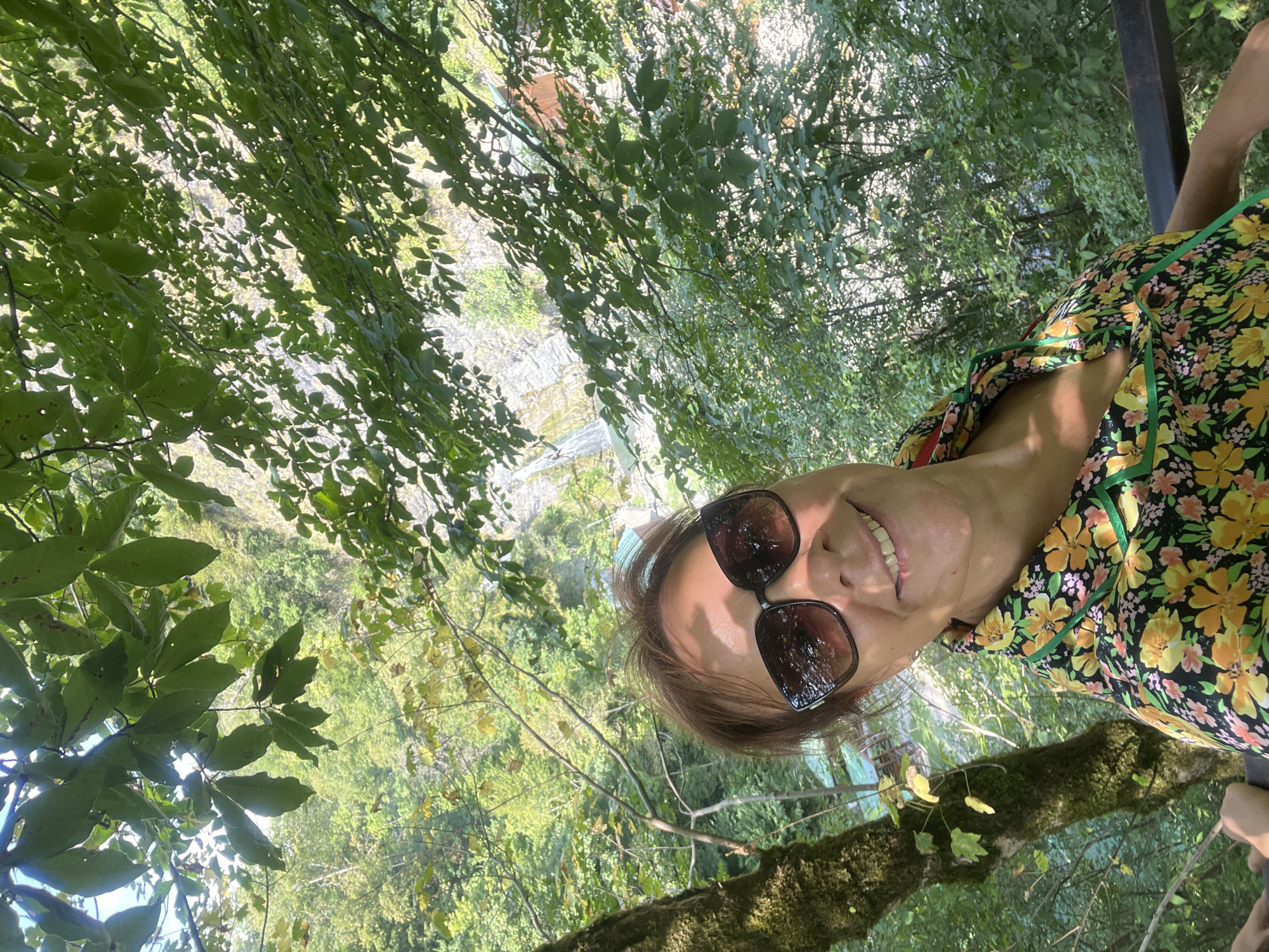 In Khostinskiy, Russia, a woman with shoulder-length brown hair and sunglasses is captured in a selfie. She is smiling and wearing a floral shirt with yellow and orange flowers on a black background and a green collar. The background suggests a natural setting with lush green trees and foliage. In the distance, a waterfall cascades down a rocky cliff face, and a body of water can be seen below. Some buildings, possibly rustic structures, are also visible amidst the greenery. A moss-covered tree trunk is to the left of the woman.