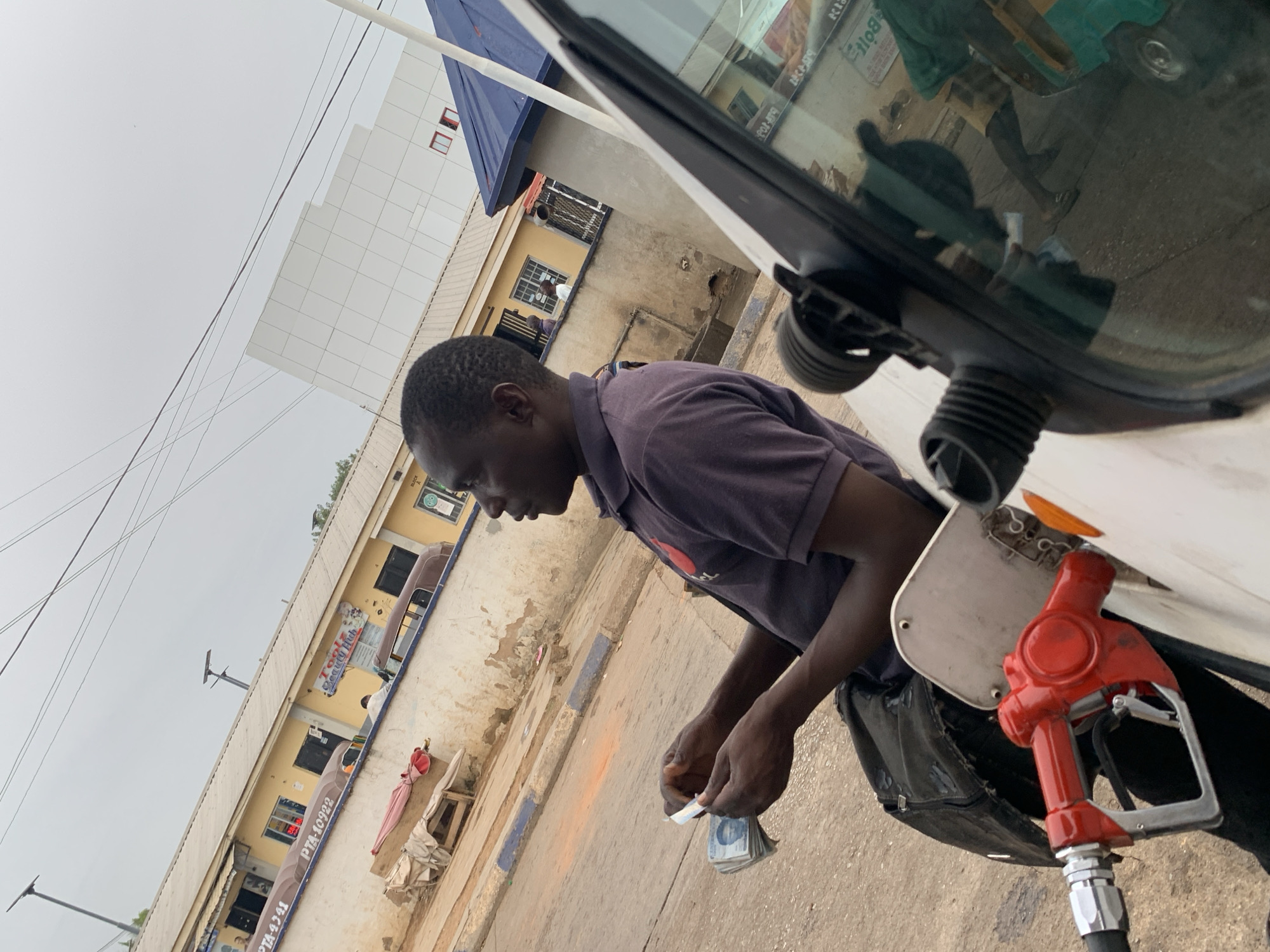 A man is pumping gas into a white vehicle. He is wearing a dark shirt and dark pants and counting currency. The gas pump is red and silver. Buildings are visible in the background, including signs for "Tools Beauty Hub" and PTA numbers. The location is likely Jos, Nigeria.