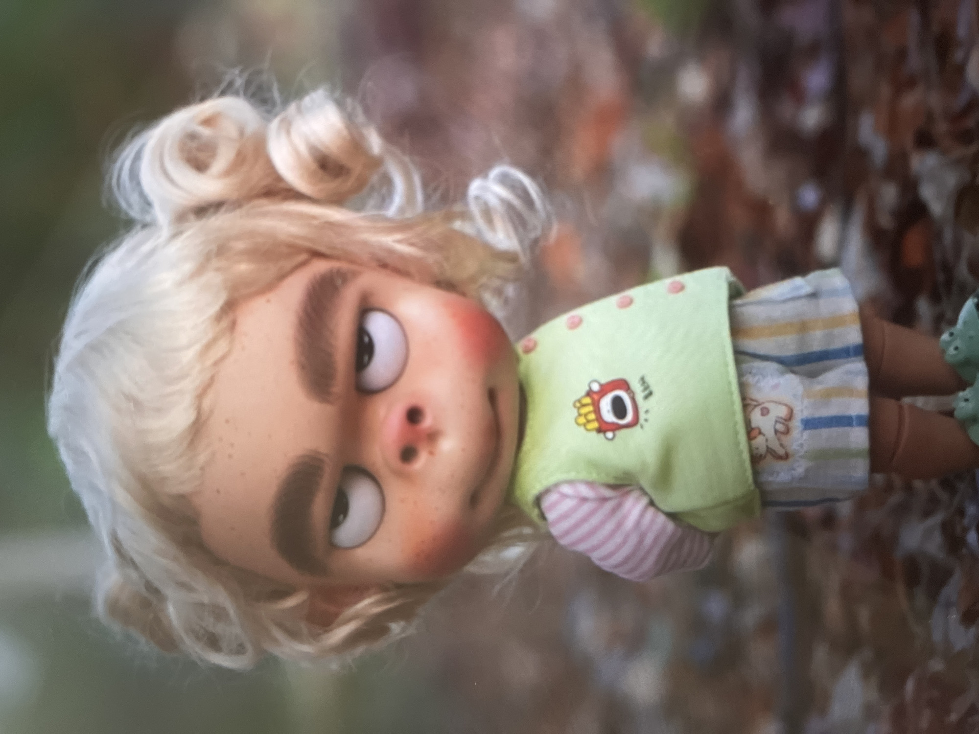 A doll with blonde curly hair, pale skin, and large eyes is standing in an outdoor setting. The doll is wearing a light green vest with a cartoon image of french fries on it, a pink and white striped long-sleeved shirt, and striped shorts. The doll's feet are clad in green shoes that resemble frogs. The background appears to be a wooded area with blurred foliage and fallen leaves. The lighting suggests it is daytime. The doll is positioned facing slightly to the left and its expression is somewhat grumpy or unimpressed. There are no other people or animals visible in the image.