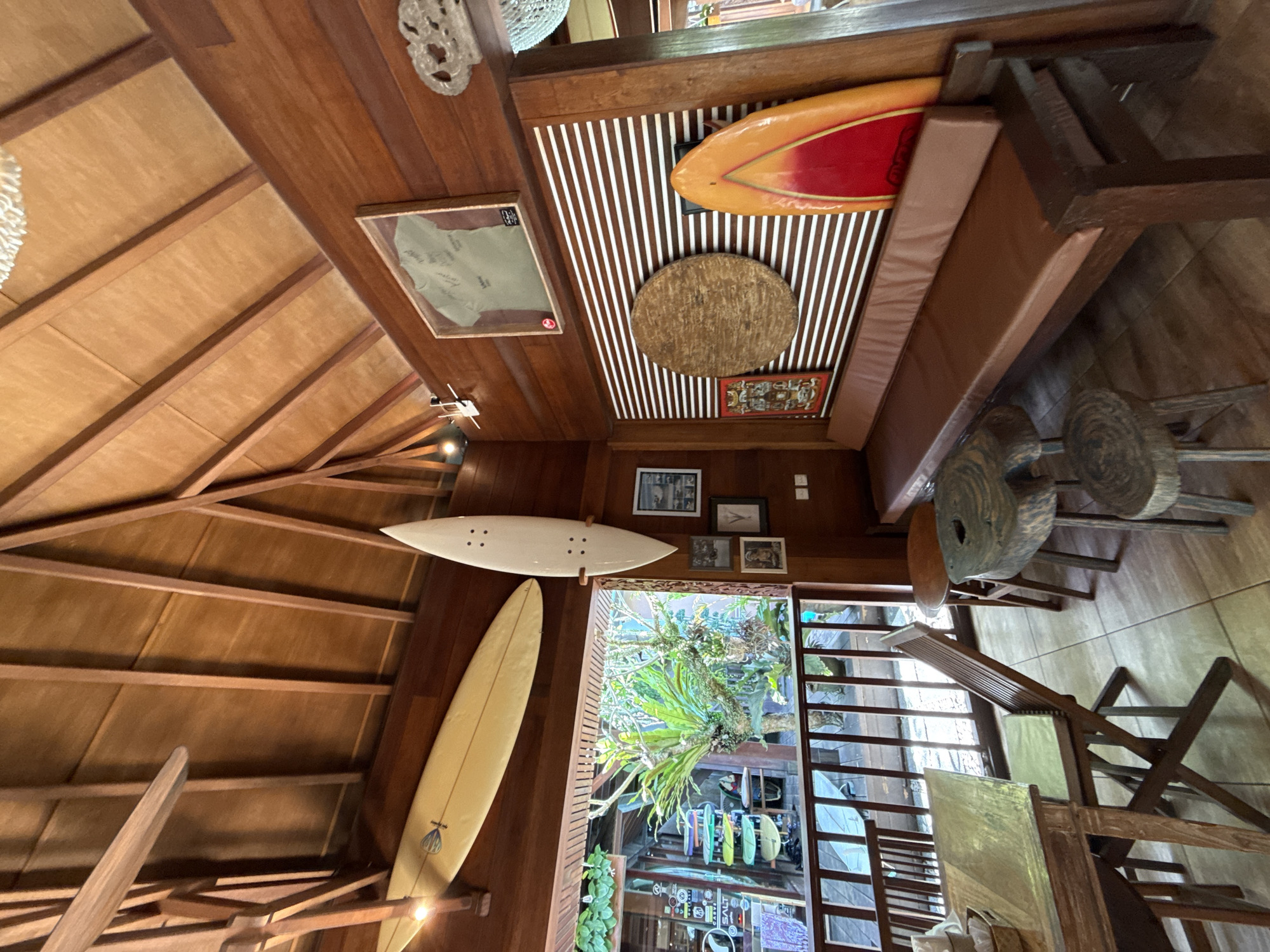 The image captures an interior space with a strong emphasis on wood construction and nautical themes. The location appears to be in a private residence. The wooden ceiling slopes upwards, supported by exposed wooden beams, and features a collection of decorative items, including two surfboards of different shapes and colors mounted on the wall. There is a framed map and a miniature airplane model also attached to the ceiling.
A built-in bench with a brown cushion lines one wall, above which is a ceiling adorned with white and brown stripes, a circular woven mat, and a framed piece of art. The walls are decorated with a series of small framed pictures.
The interior opens up to an outdoor area through a glass enclosure with vertical slats. The exterior features lush greenery and a multi-level display of various surfboards. The furniture in the interior includes a series of wooden stools, and a wooden folding chair.