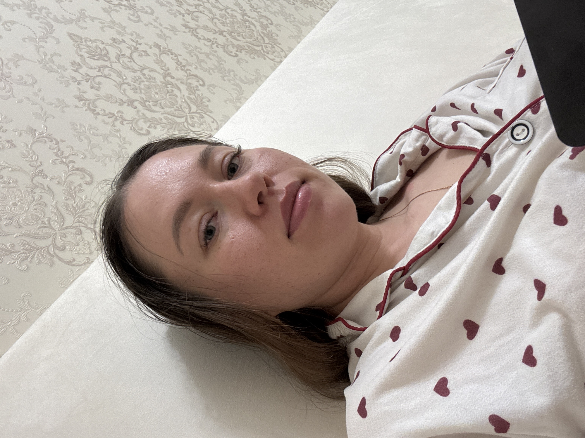A woman with light skin, brown hair, and blue eyes is lying on a white surface, possibly a bed, wearing a white pajama top with small red hearts. The background includes a beige wall with a floral pattern. Part of a dark object, possibly a phone or tablet, is visible on the right side of the image. The location is Ферма, Russia.