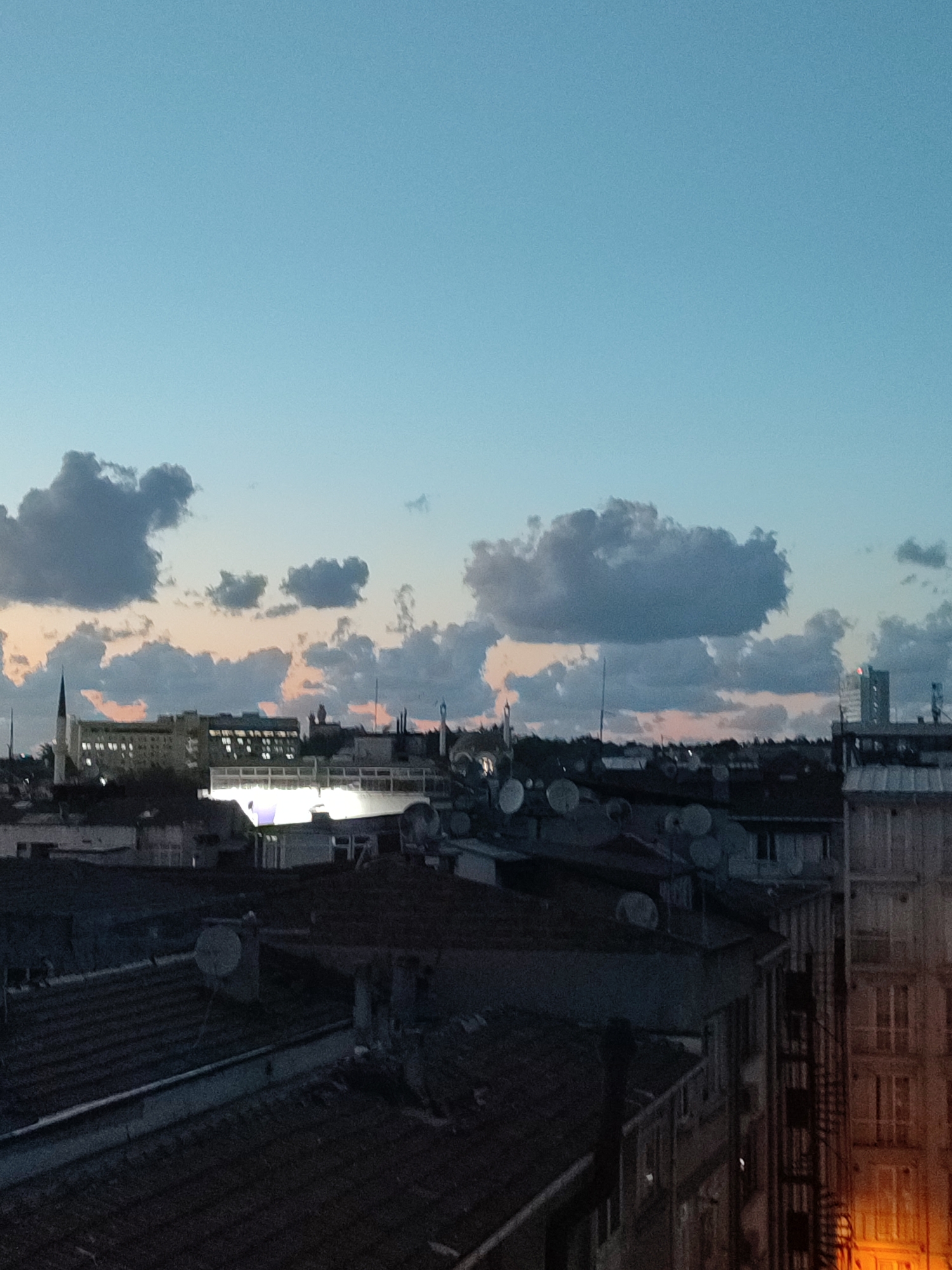 This is a twilight aerial view of a dense urban environment, likely Kadıköy, Turkey. The sky is a gradient of pale blue, transitioning to soft hues of orange and pink near the horizon, indicating either sunrise or sunset. Fluffy, dark gray clouds are scattered across the sky, some casting shadows on the city below.
The foreground and mid-ground are dominated by a dense cluster of buildings. Numerous rooftops are visible, many covered with dark tiles. Several satellite dishes are prominent on some of the roofs. A tall, spire-like structure is visible on the left side of the image, possibly a church steeple or a minaret. In the mid-ground, a large, multi-story building with many illuminated windows stands out, suggesting it is an office or residential complex. Further to the right, another modern high-rise building can be seen.
The overall impression is of a bustling city at a transitionary time of day. No people are discernible in the image. The scene is still and quiet, capturing the architectural landscape under the soft light of twilight.