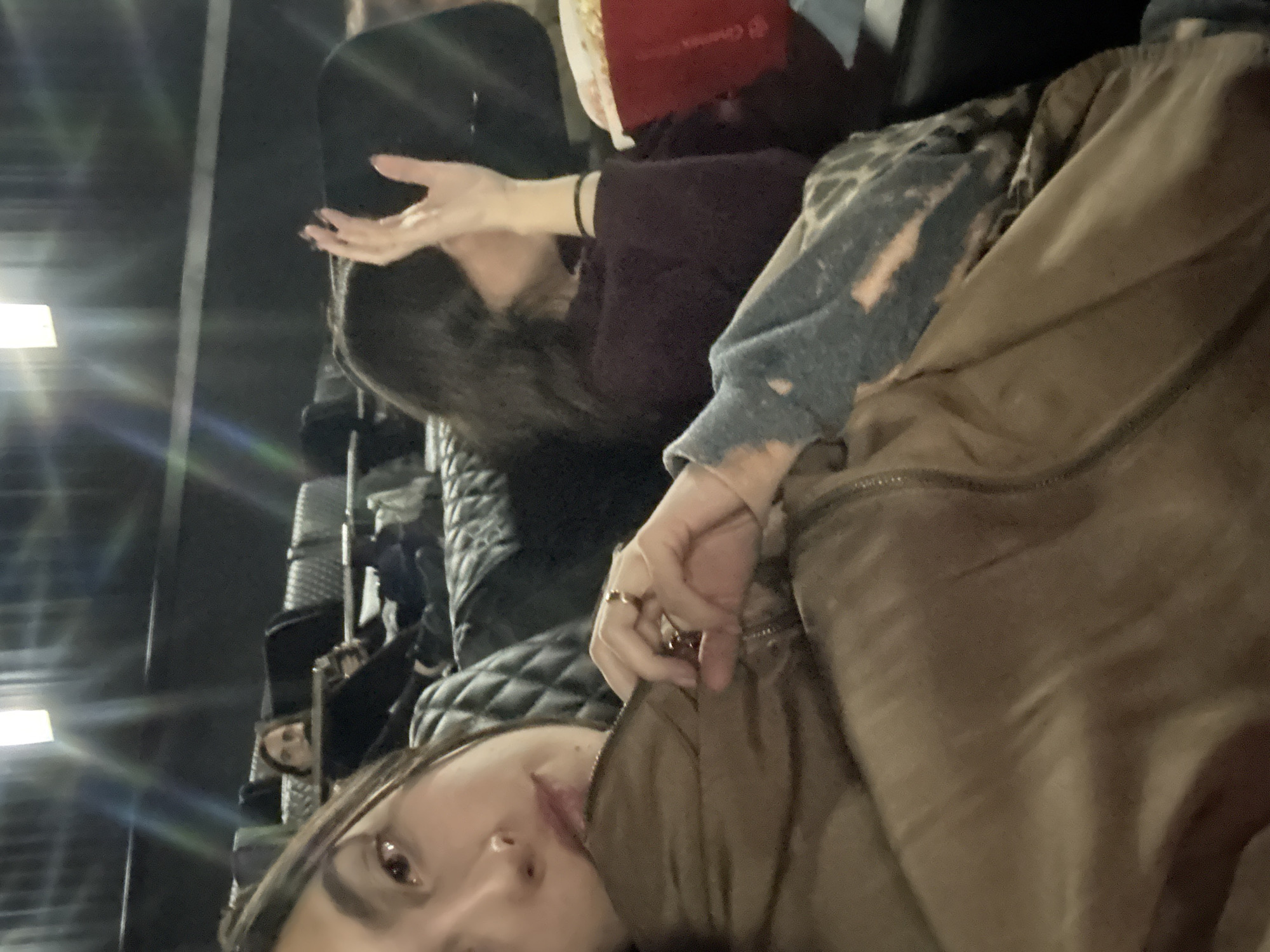 The image appears to be a selfie taken by a person sitting in what seems to be a movie theater, possibly in Mexico City, Mexico (due to the presence of "Cinépolis" branding, a Mexican cinema chain). The person taking the selfie is in the foreground, with part of their face visible near the bottom of the frame. They are wearing a brown jacket. To the left, several theater seats can be seen.
Another person is in the background, sitting in one of the theater seats. This person is partially obscured, but it is possible to see the hand and head of the person. There is a box of popcorn visible and their hair is black.