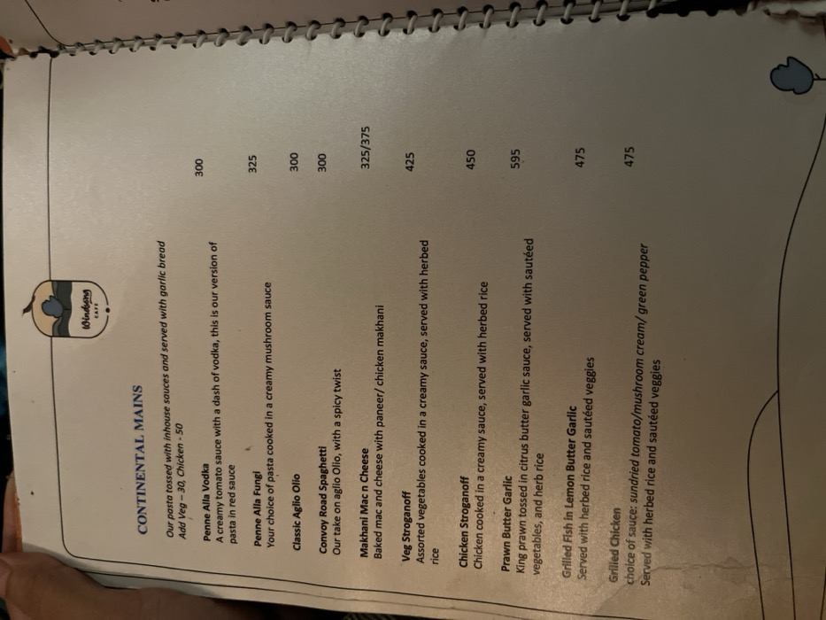 This image displays a menu from "Windsong Cafe," likely located in Dibrugarh, India. A human hand is partially visible at the bottom left, holding the spiral-bound menu open. The menu focuses on "CONTINENTAL MAINS," listing various dishes with their descriptions and prices.
The subjects on the menu include several pasta dishes like Penne Alla Vodka, Penne Alla Fungi, Classic Aglio Olio, and Convoy Road Spaghetti, priced between 300 and 325. Other main courses include Makhani Mac n Cheese (325/375, with paneer or chicken makhani options), Veg Stroganoff (425), and Chicken Stroganoff (450), both served with herbed rice. Seafood options feature Prawn Butter Garlic (595) and Grilled Fish in Lemon Butter Garlic (475), accompanied by sautéed vegetables and herbed rice. A Grilled Chicken dish (475) offers a choice of sundried tomato, mushroom cream, or green pepper sauce, also served with herbed rice and sautéed veggies. Additional costs for adding veg or chicken to pasta are listed as 30 and 50 respectively.
The scene depicts a typical restaurant or cafe setting where a patron might be selecting their meal. The menu itself is printed on white paper with dark blue text for headings and item names, and black text for descriptions. A small blue logo with "Windsong CAFE" is prominently displayed at the top left. The image is taken at an angle, suggesting it was captured handheld.