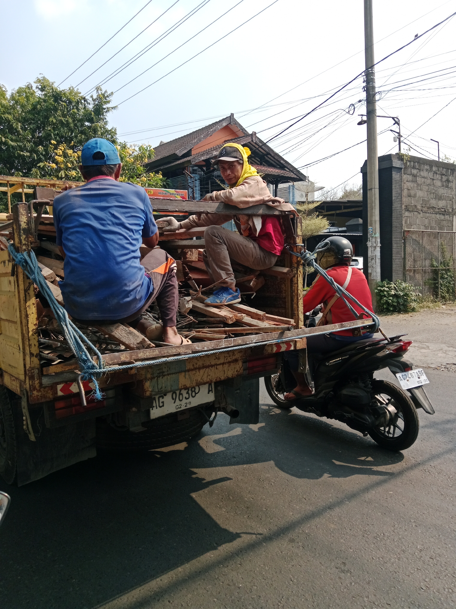 In Kediri City, Indonesia, two individuals are seated in the back of a yellow truck filled with wooden debris. One person, wearing a blue shirt and blue cap, is facing away from the camera. The other person, wearing a brown jacket and yellow head covering, is facing the camera and resting their arm on the truck's railing. A blue rope is tied to the side of the truck, securing the debris. In the foreground, a person wearing a red shirt and black helmet is riding a black motorcycle, with the truck's rear license plate visible, reading "AG 9638 A". The scene appears to be on a paved road with a brick wall and buildings in the background.