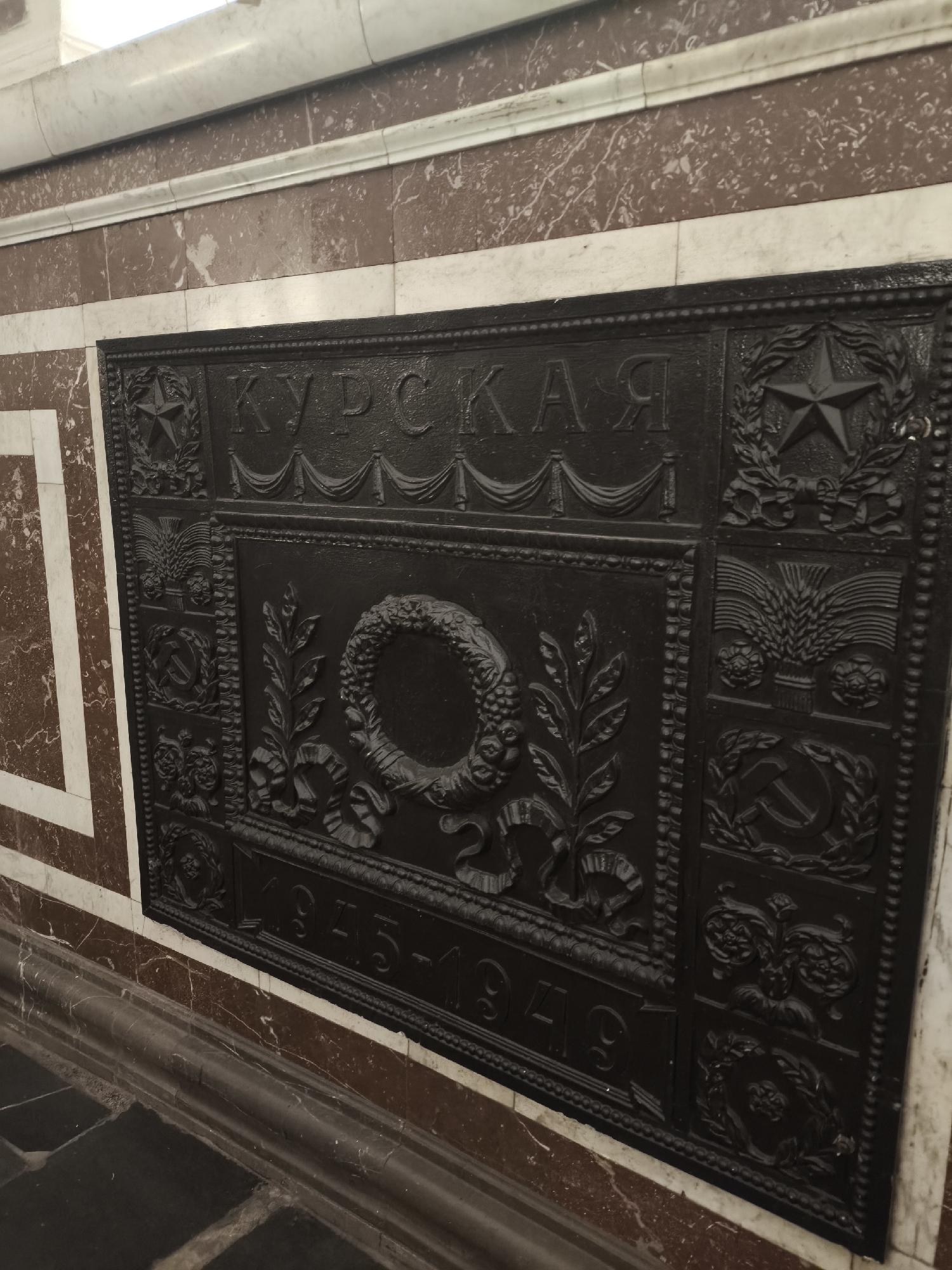 The image shows a decorative panel featuring Soviet symbolism in a building, possibly a metro station. The panel is located in an unknown city.
The panel is made of a dark-colored material, possibly metal, and has a raised design. The text "КУРСКАЯ" (Kurskaya) is prominently displayed at the top. Below the text is a frame containing a wreath design. Beneath this wreath, the date "1945-1949" is inscribed. The panel also incorporates communist symbols such as a five-pointed star, a hammer and sickle, and depictions of wheat.
The panel is affixed to a wall with a two-tone color scheme of a reddish-brown and white marble-like material, with the marble covering the floor. The lighting in the area appears to be relatively dim.