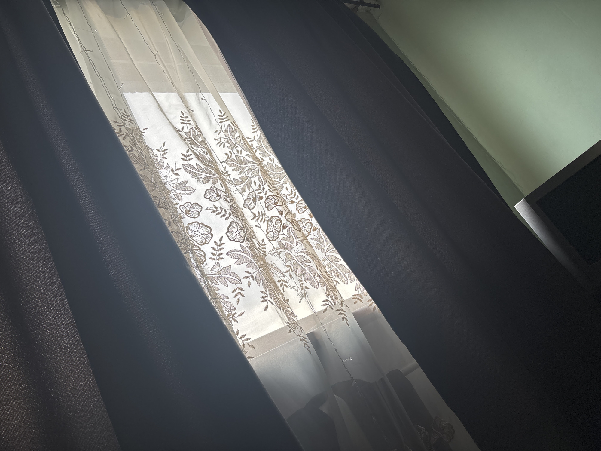 The image shows a window with two layers of curtains. The inner layer is a sheer, light-colored curtain with a floral pattern. The outer layer is a dark, heavy curtain. Light is filtering through the gap between the curtains. A small portion of a green wall and a dark object (possibly a TV) are visible in the background. Location is unknown, Russia.