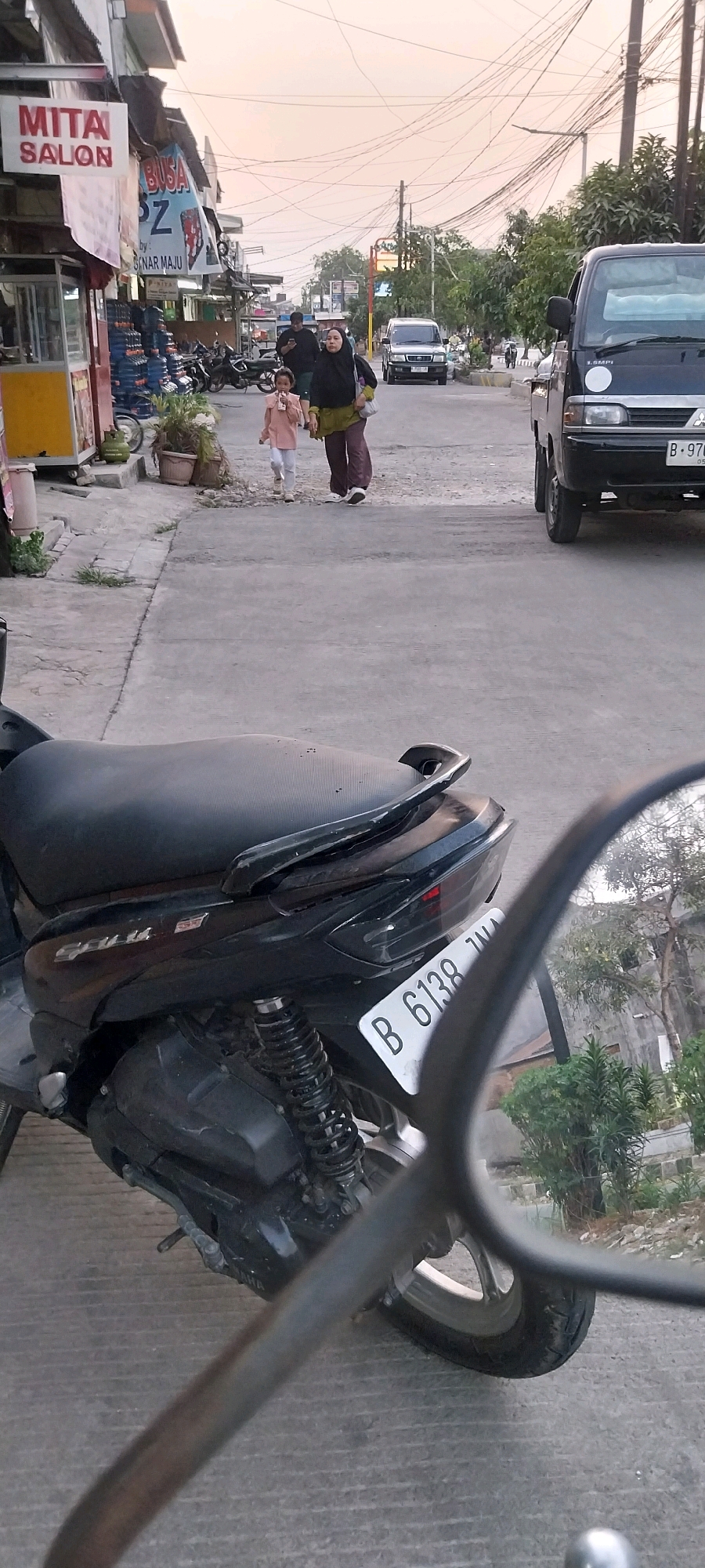 In Bojong Nangka, Indonesia, a street scene unfolds with a close-up of a black motorcycle in the foreground, its license plate displaying "B 6138 JNA". To the left, a roadside shop with a sign reading "MITA SALON" is visible, featuring a yellow food stall and stacked water jugs. Further down the street, two individuals, a woman in a headscarf and a child, walk away from the camera. The woman wears a dark headscarf and a green top with dark pants, while the child is dressed in a light pink outfit. Behind them, a black pickup truck with a license plate starting with "B 976" is parked on the road. The street is lined with trees and utility poles with a complex network of overhead wires, suggesting an urban or semi-urban environment. The sky is a pale, diffused orange, indicating either sunrise or sunset.