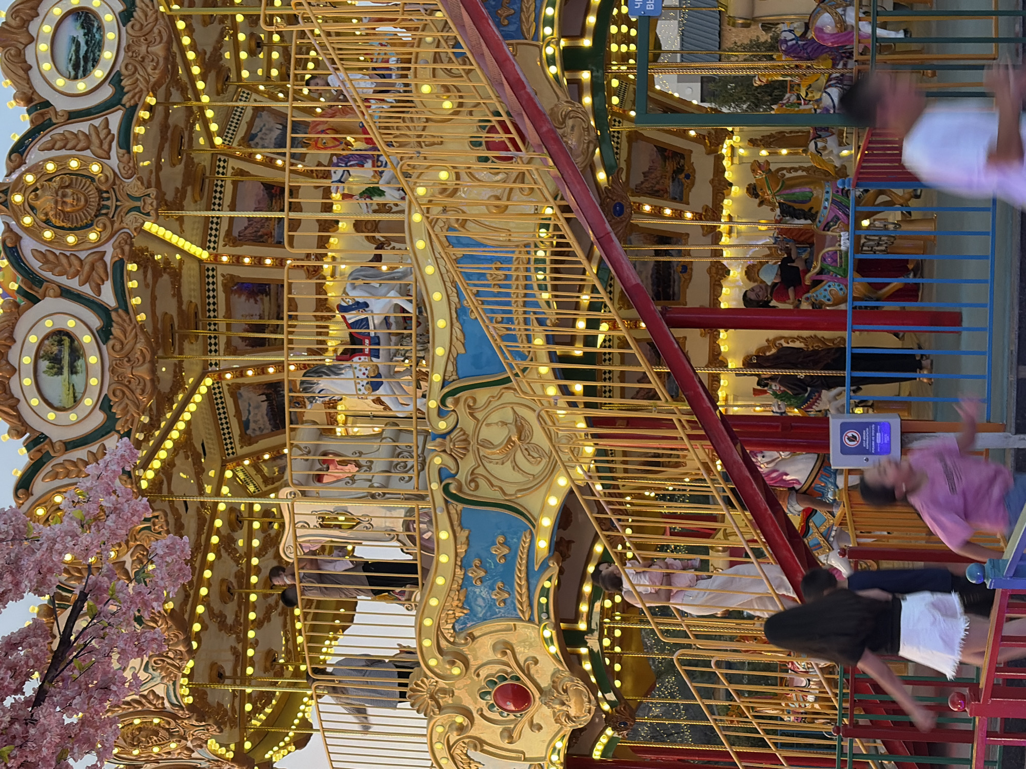 A carousel in an amusement park is pictured from a low angle. The carousel is illuminated by numerous small, yellow lights and decorated with ornate gold trim and painted panels. Pink artificial cherry blossoms hang from the top left of the frame. Several people are visible on and around the carousel. On the carousel's upper level, a person holds a baby. On the stairs leading to the upper level, a person carries a child. In the foreground, several children and adults are standing behind a colorful fence. One child in a pink shirt is in motion. The carousel appears to be in operation. The background is blurred but suggests a city environment with lights.