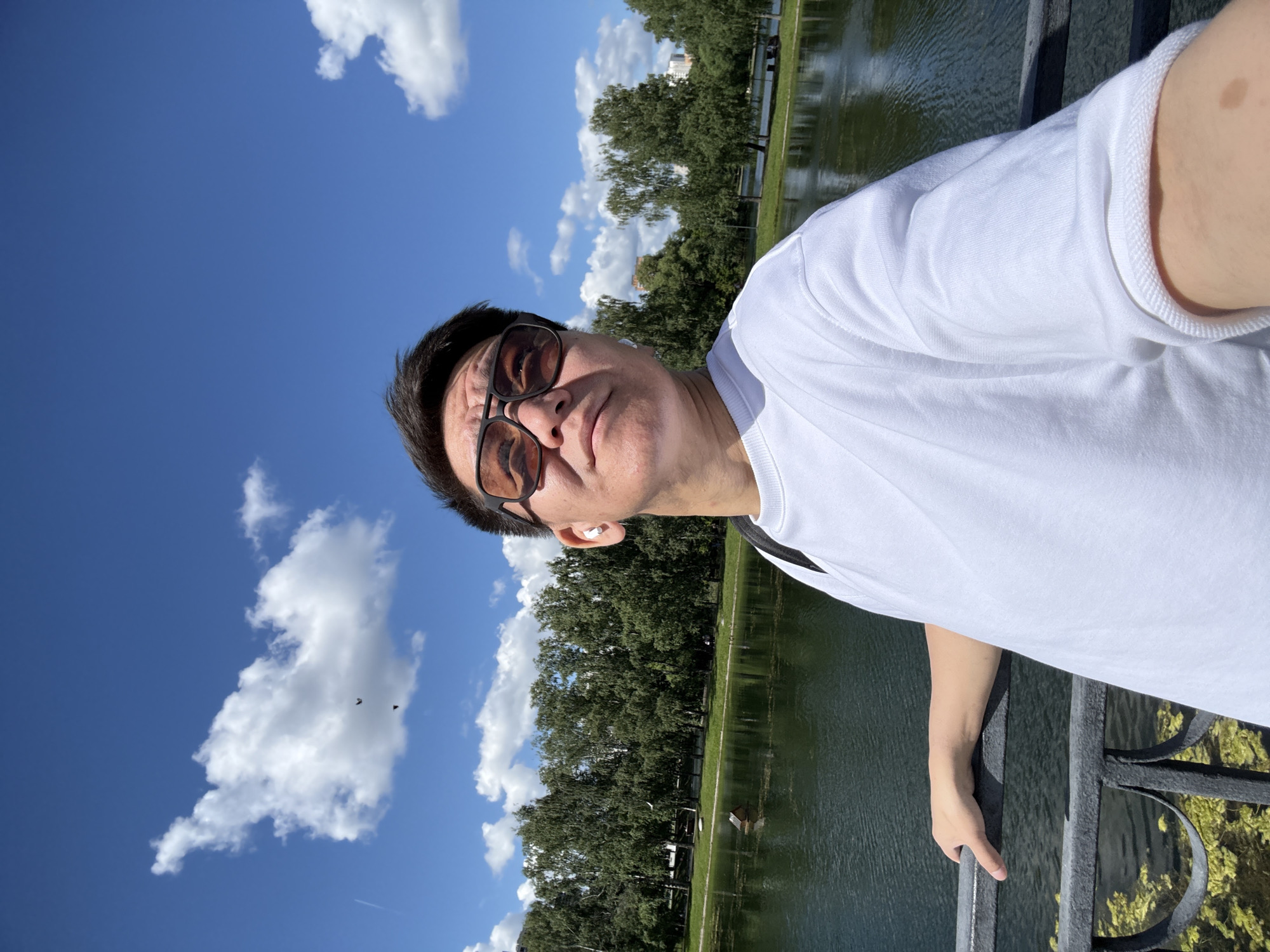 A man stands on a bridge, leaning on the railing, taking a selfie. He is wearing a white t-shirt and sunglasses. In the background, there is a body of water with trees and buildings lining the shore. The sky is blue with scattered clouds. The location is identified as Moscow, Russia.