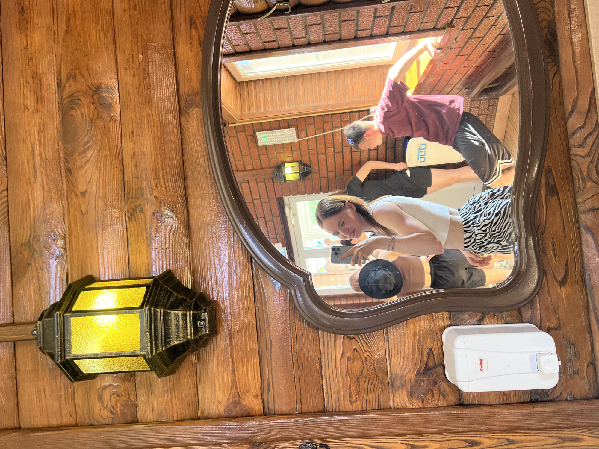 The image is a reflection in an ornate mirror mounted on a wood-paneled wall. The mirror reflects an interior space where three people are visible: a young woman holding a smartphone, presumably taking the picture; a man standing on a raised surface; and another person partially visible in the background. The environment in the reflection includes a skylight, brickwork, and what appears to be a light fixture. The wall on which the mirror is mounted features an antique-style light fixture and a white soap dispenser. The location is Grishanki, Russia.