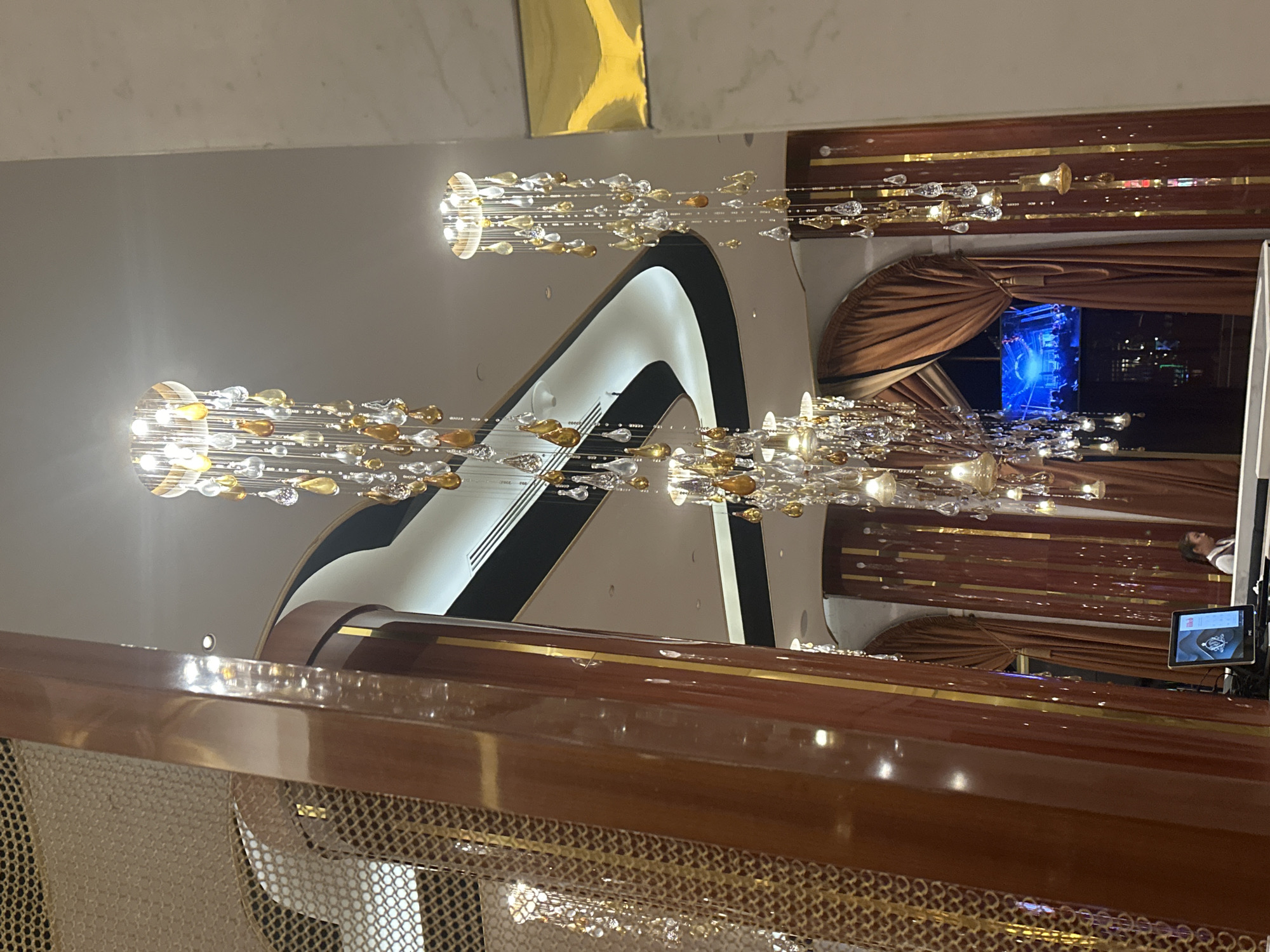 This image depicts an opulent interior, likely a hotel lobby or a high-end establishment, in Estosadok, Russia. The scene is dominated by dramatic, cascading chandeliers with crystal and amber-colored teardrop accents, hanging from a high, modern ceiling with curved architectural elements.
In the foreground, a large, polished wooden column with golden trim is prominent. To the left, a metallic, mesh-like screen adds a decorative element. In the mid-ground, rich, rust-colored drapes frame an archway. Through this archway, a television screen displays a bright blue and white concert scene.
At the reception desk, partially obscured by the drapes and chandeliers, a woman in a white shirt and dark vest is visible, appearing to be a staff member. A computer monitor sits on the desk in front of her. The overall impression is one of luxury and sophistication, with a blend of classic and contemporary design elements.