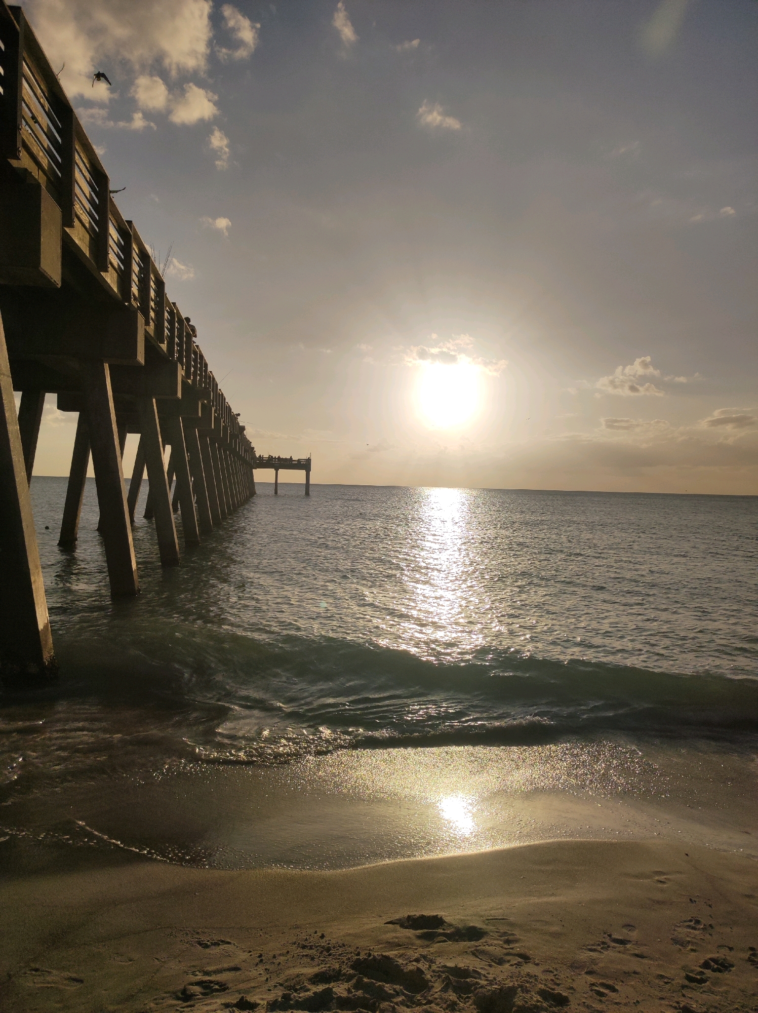 The image depicts a view from a beach in Venice, United States of America, looking towards a pier extending into the ocean at sunset. The sun is low on the horizon, casting a warm glow and reflecting off the water's surface. Waves are gently breaking on the sandy shore. The pier, with its wooden railings and concrete pilings, stretches out into the distance, with several people visible fishing from its structure. Two birds are in flight in the sky above the pier. The sky is partly cloudy, with the sun partially obscured by a cloud. The overall scene is serene and evokes a sense of calm.