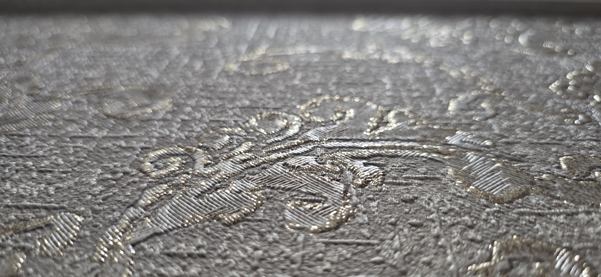 The image displays a close-up, macro view of a textured surface, likely wallpaper or fabric, with a repeating damask-like pattern. The primary color of the surface is a muted, textured gray, while the pattern elements are highlighted with a metallic gold or silver thread, catching the light and creating a subtle shimmer. The pattern itself is ornate, featuring swirling motifs and linear textures within the design. The lighting is soft and directional, creating highlights and shadows that emphasize the three-dimensionality of the texture. The background is out of focus, suggesting a shallow depth of field, and appears to be a continuation of the same patterned surface, fading into a softer, bokeh effect. There are no people, animals, or identifiable objects from a cityscape visible in this close-up. The image does not provide any information that would allow for its location to be determined within the city of Urupskiy, Russia, or any other specific location.