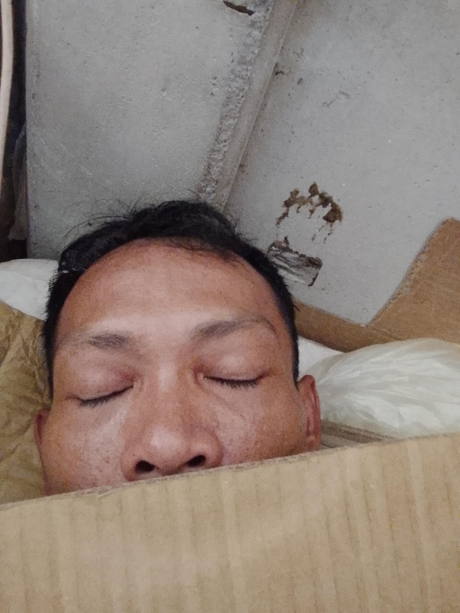 The image shows the face of a person, likely male, with eyes closed, suggesting sleep or relaxation. The person is partially covered by a tan, corrugated cardboard object, obscuring the lower portion of the face and body. The individual has dark hair. The background consists of a gray concrete or plastered wall with visible imperfections and stains. The upper left corner shows a portion of what appears to be white cloth or curtains. To the person's right is a partially visible white pillow. The location is specified as Bukit Ketok, Indonesia.